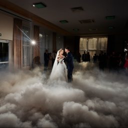 FOTOFRAJDA ALEKSANDRA MICEK - Para młoda tańcząca pierwszy taniec w chmurze dymu na sali weselnej, w tle goście i reflektor.