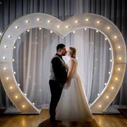 FOTOFRAJDA ALEKSANDRA MICEK - Para młoda w Rzeszowie całująca się na tle białego, podświetlanego serca z żarówkami, ustawionego na drewnianej podłodze przed białymi, prześwitującymi zasłonami.