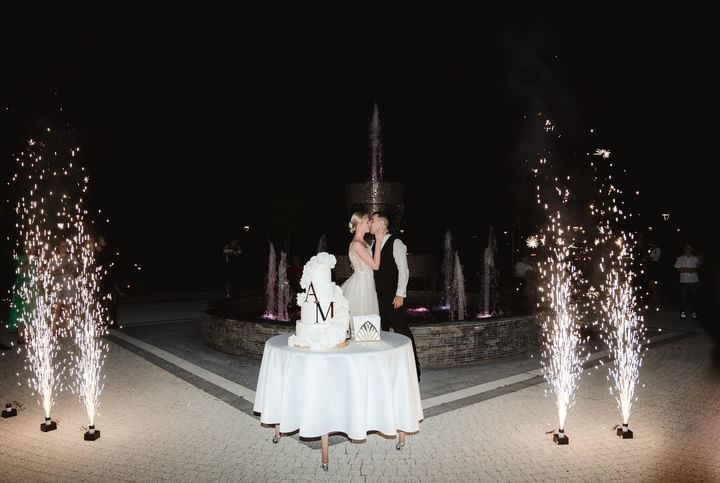 Pocałunek pary młodej przy torcie weselnym z inicjałami i fontannami iskier na tle fontanny w nocy.