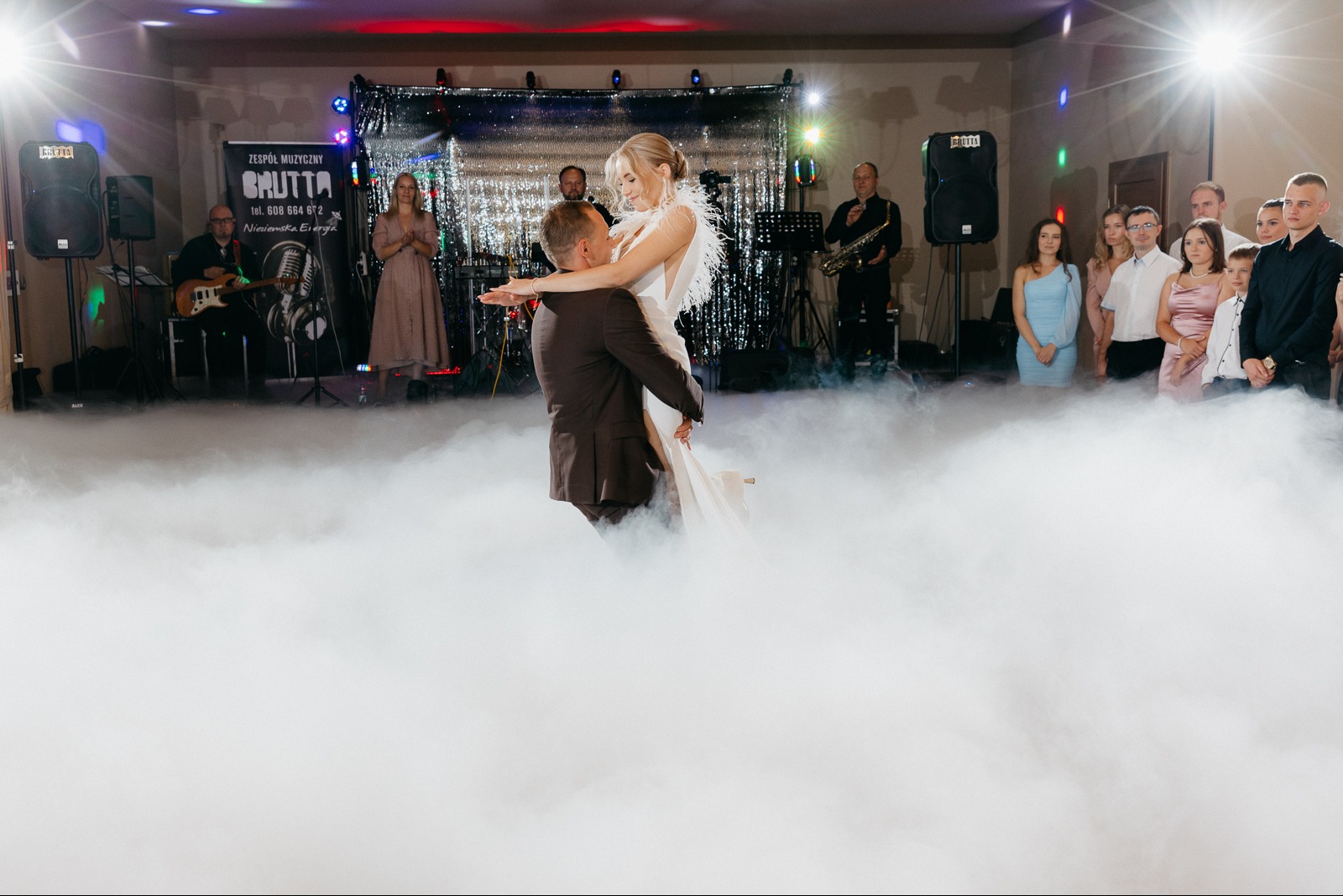 Para młoda w tańcu unoszona przez pana młodego na tle zespołu muzycznego i gości, efekt ciężkiego dymu na parkiecie, oświetlenie sceniczne.