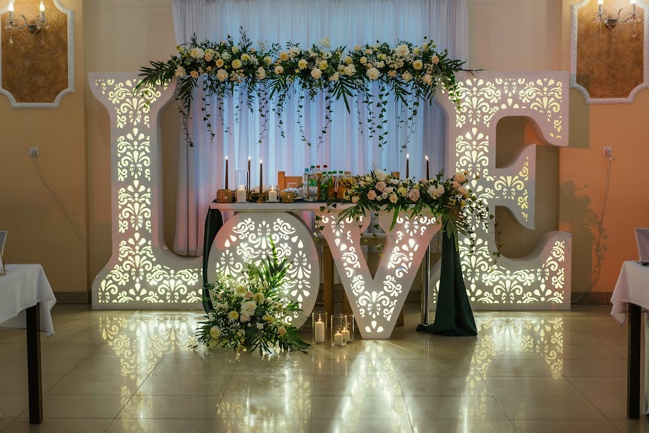 Dekoracja sali weselnej z podświetlanymi literami LOVE, kwiatami i świecami, ustawiona przed zasłoniętym oknem.