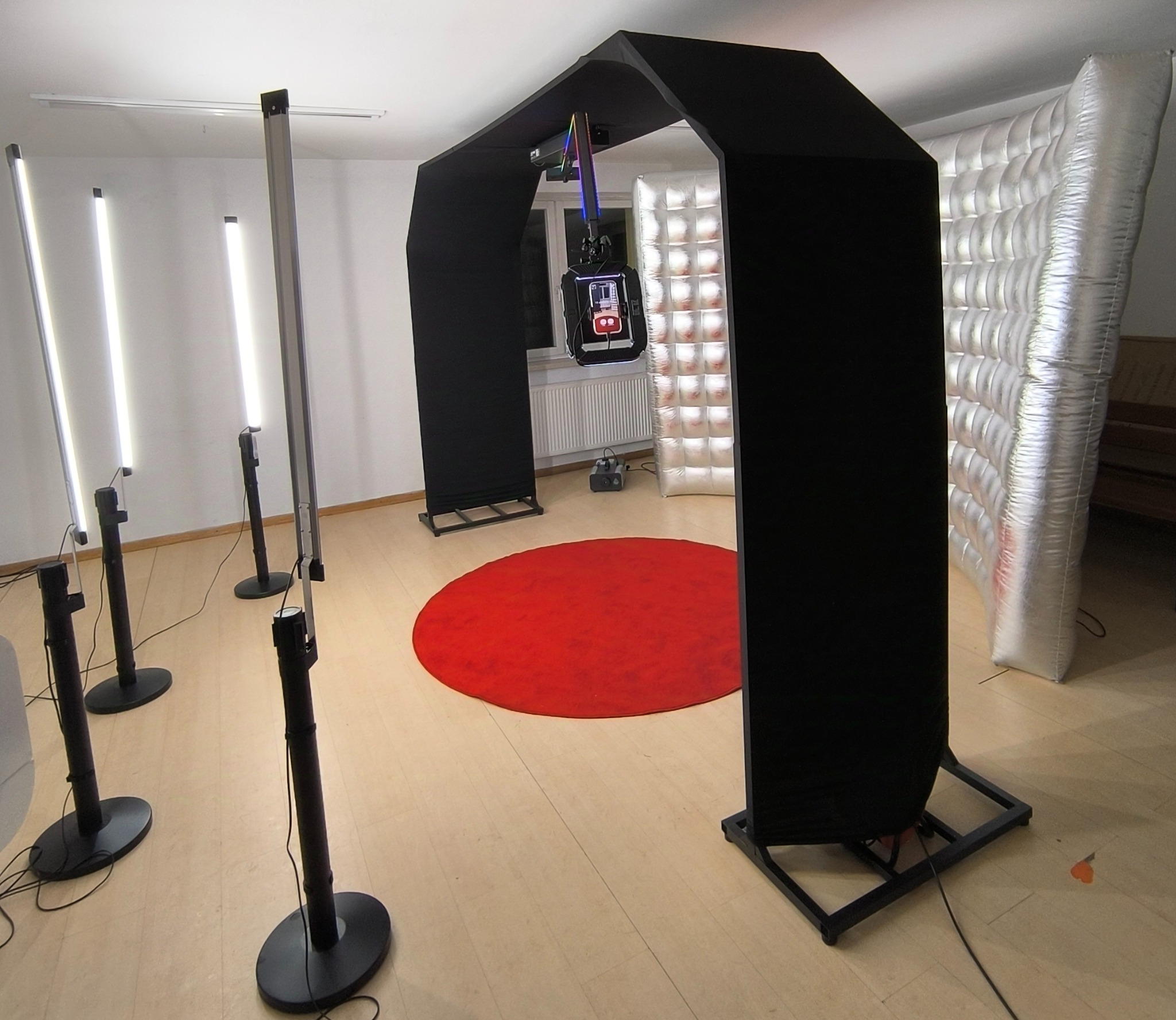 Wnętrze z rozstawioną czarną fotobudką z ekranem, oświetleniem LED i dmuchanym tłem, czerwony dywan na podłodze.