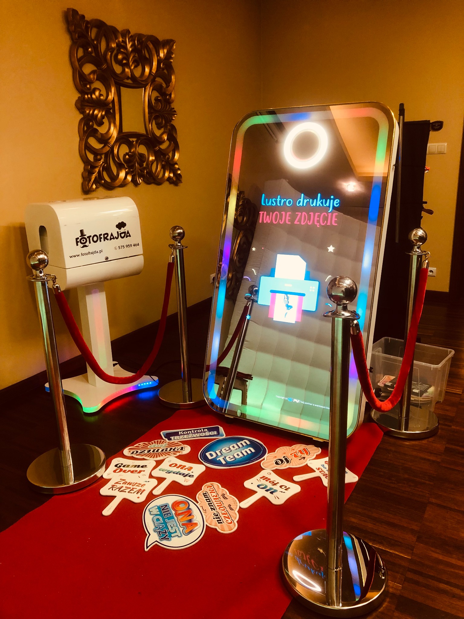 Nowoczesna fotobudka lustrzana z oświetleniem LED i napisem 'Lustro drukuje TWOJE ZDJĘCIE', otoczona słupkami z czerwonymi sznurami, obok biała drukarka do zdjęć i rekwizyty na czerwonym dywanie.
