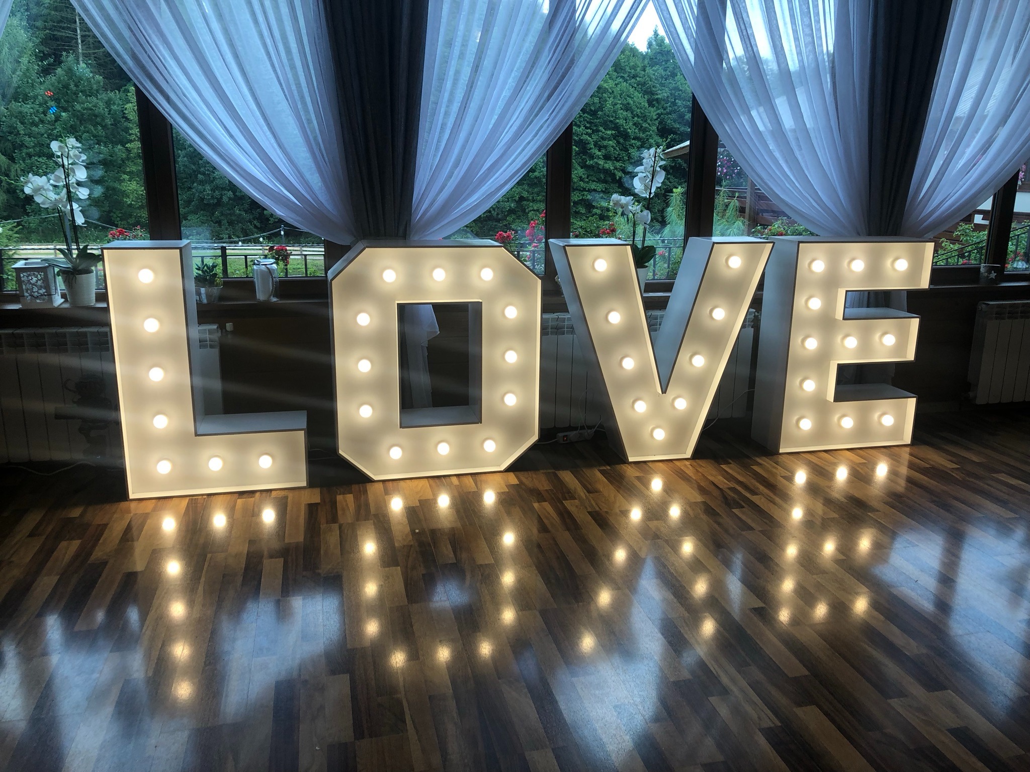 Świecący napis LOVE na parkiecie, duże litery z żarówkami, w tle okno z firankami i zielenią