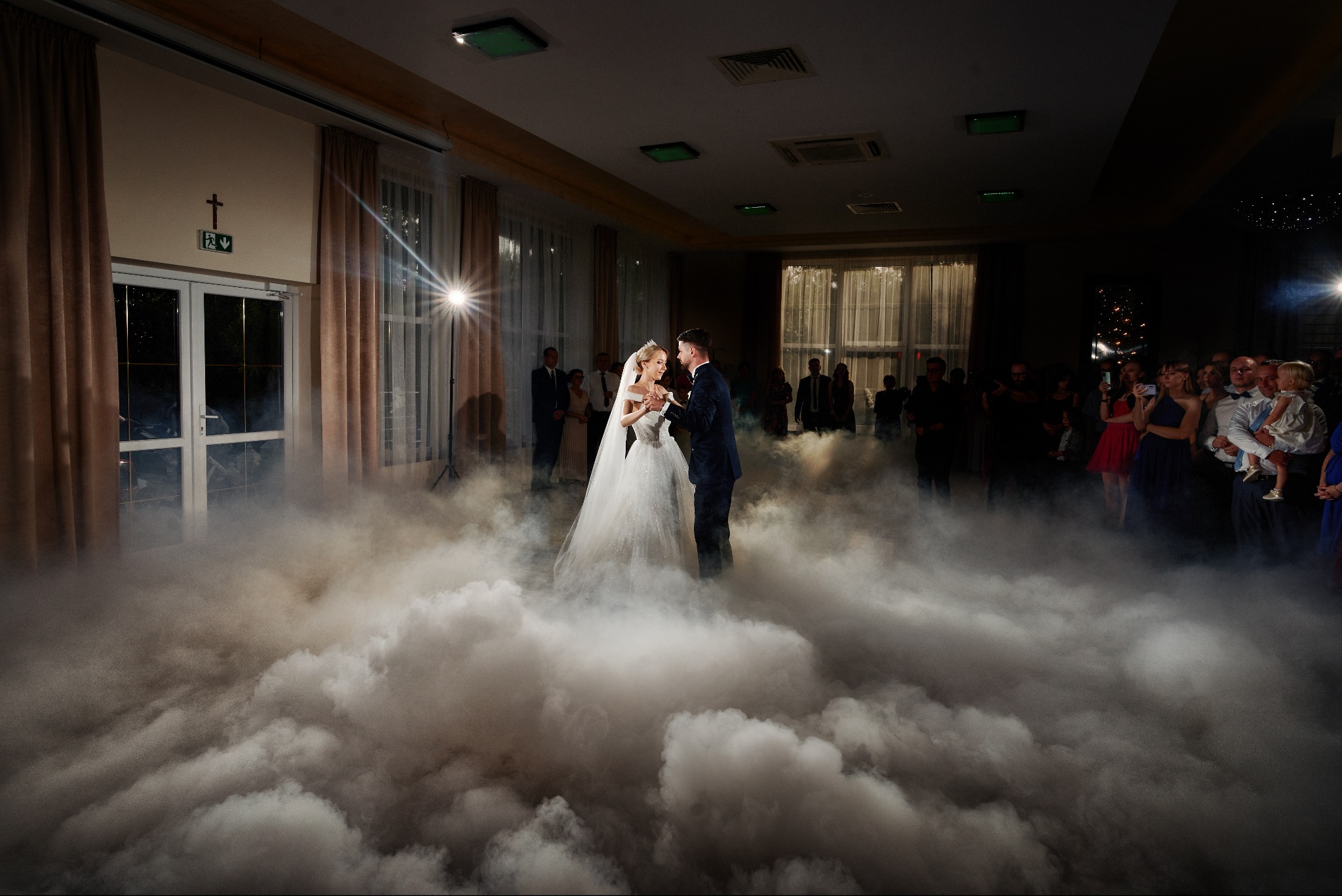 Para młoda tańcząca pierwszy taniec w chmurze dymu na sali weselnej, w tle goście i reflektor.