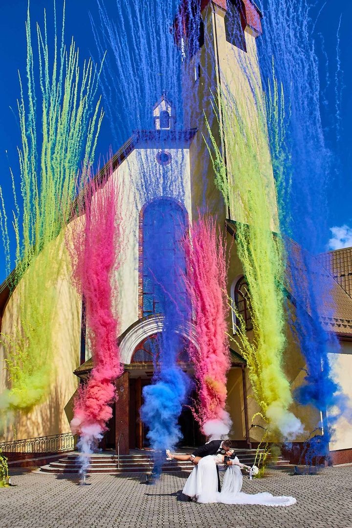 Para młoda przed kościołem, otoczona kolorowymi strumieniami dymu, tworzącymi efektowną oprawę uroczystości.