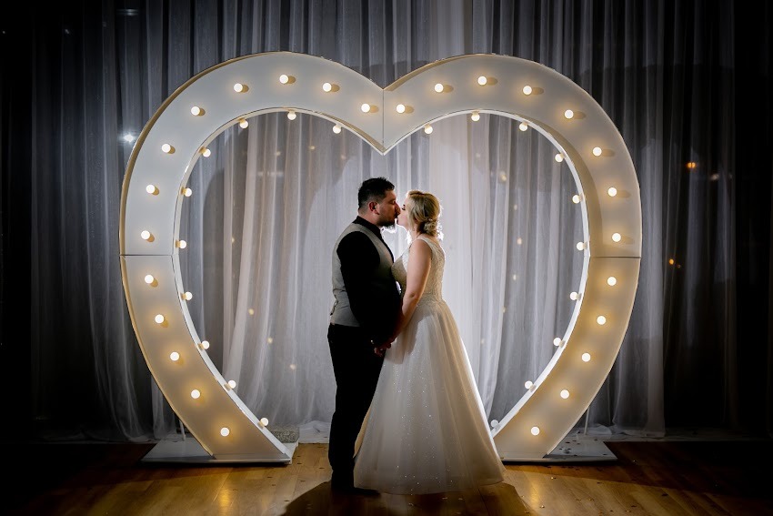 Para młoda w Rzeszowie całująca się na tle białego, podświetlanego serca z żarówkami, ustawionego na drewnianej podłodze przed białymi, prześwitującymi zasłonami.