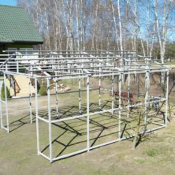 Stalowa konstrukcja szkieletowa przyszłej altany ogrodowej, w tle dom z zielonym dachem i brzozowy lasek, słoneczny dzień, cień konstrukcji na trawie.