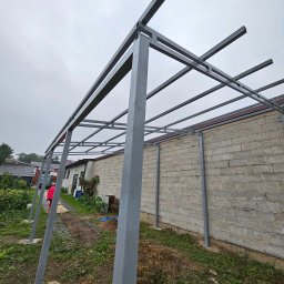 Formithal - Szara, metalowa konstrukcja z profili kwadratowych, prawdopodobnie zadaszenie, w trakcie budowy. Widoczny fragment budynku z bloczków betonowych i niebo.