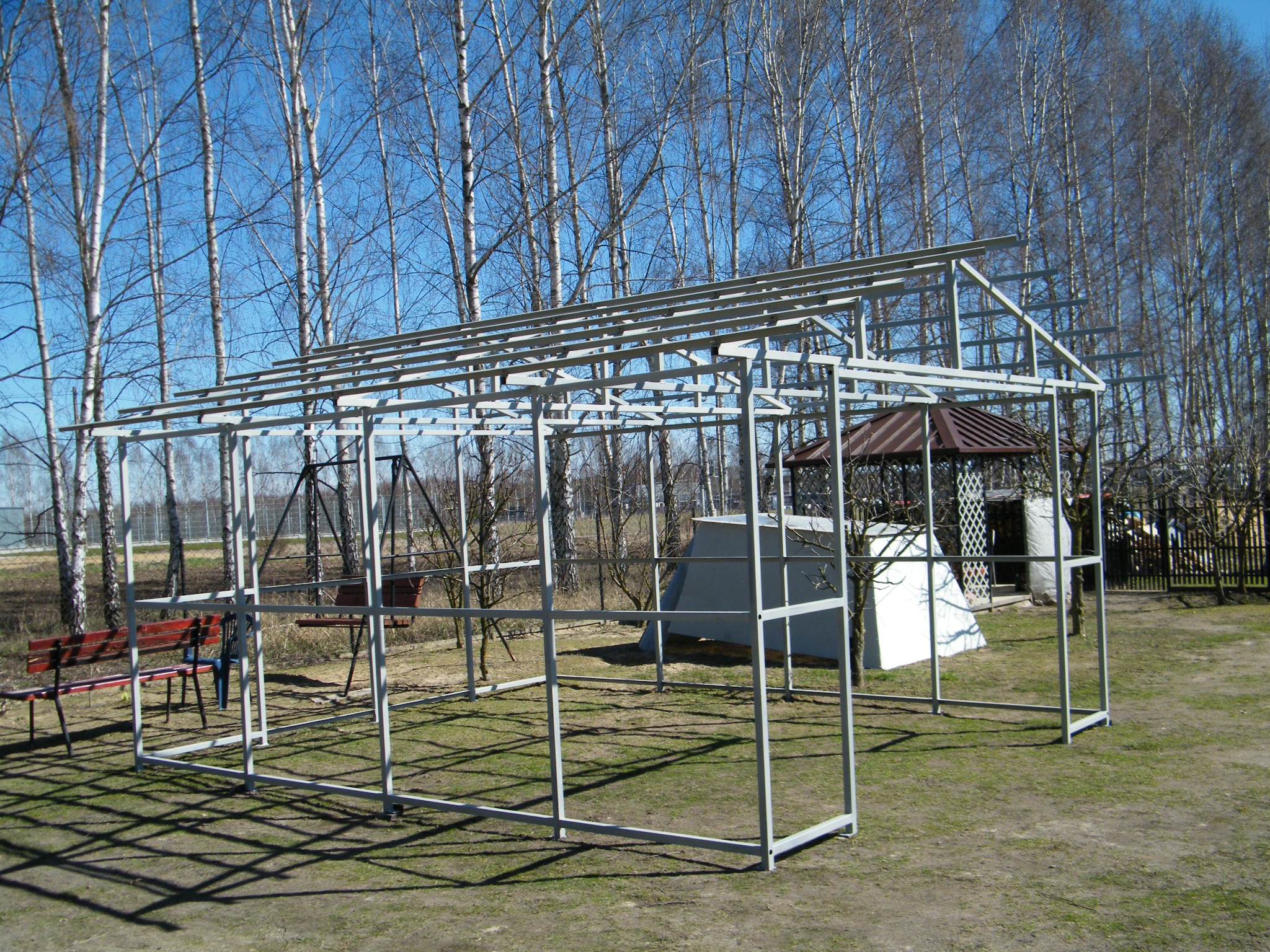 Metalowa konstrukcja szkieletowa na trawniku, prawdopodobnie pod budowę wiaty lub szklarni, z widocznym zarysem dachu i ścian. W tle drzewa i altana.