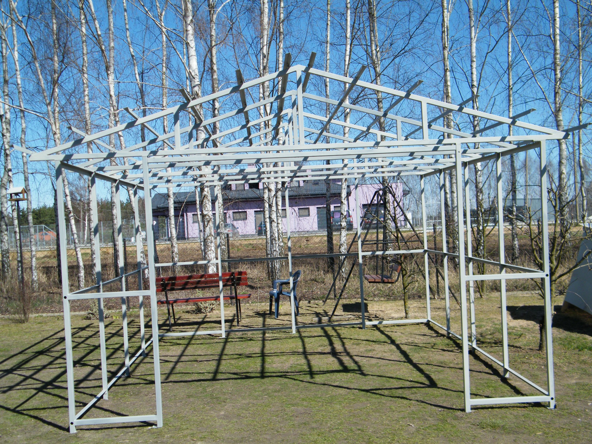 Szkielet metalowej konstrukcji altany ogrodowej, z widocznymi elementami ławek i huśtawki wewnątrz, na tle drzew i budynku mieszkalnego w oddali.