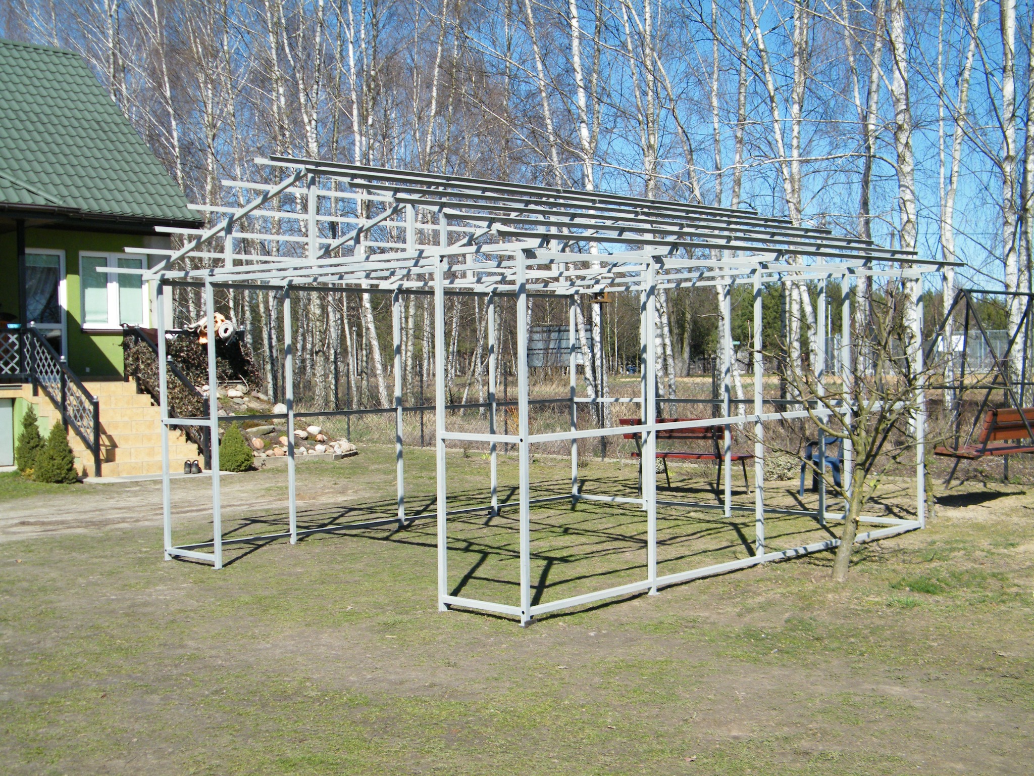 Metalowa konstrukcja szkieletowa, prawdopodobnie altany lub pergoli, na trawiastym terenie, z drzewami w tle, w słoneczny dzień. Widoczne elementy spawane.