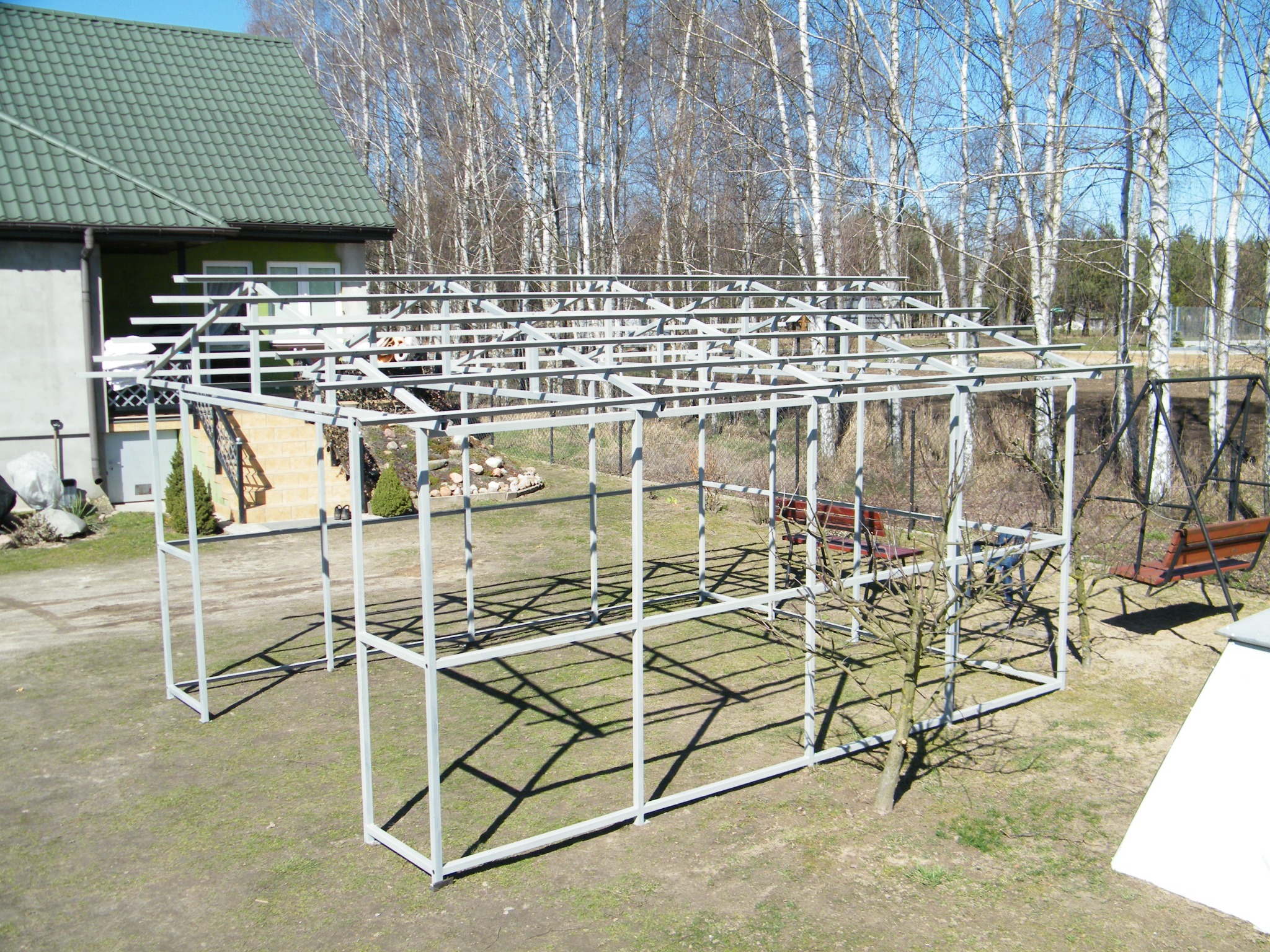 Stalowa konstrukcja szkieletowa przyszłej altany ogrodowej, w tle dom z zielonym dachem i brzozowy lasek, słoneczny dzień, cień konstrukcji na trawie.