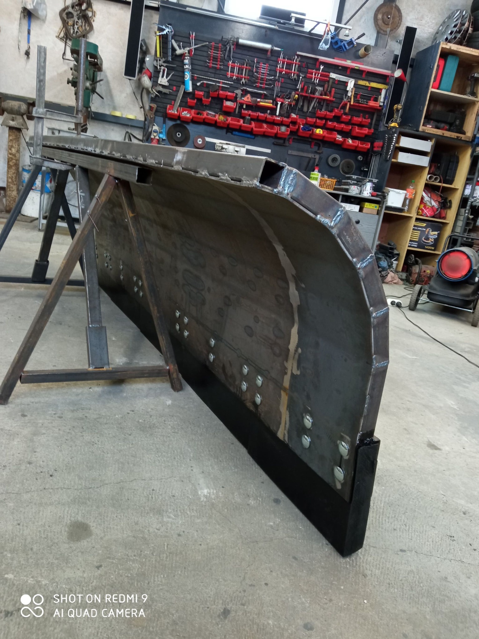 Metalowy pług śnieżny w warsztacie, spawane elementy, widoczne ślady obróbki. Pług stoi na metalowym stelażu, w tle narzędzia i regały warsztatowe.