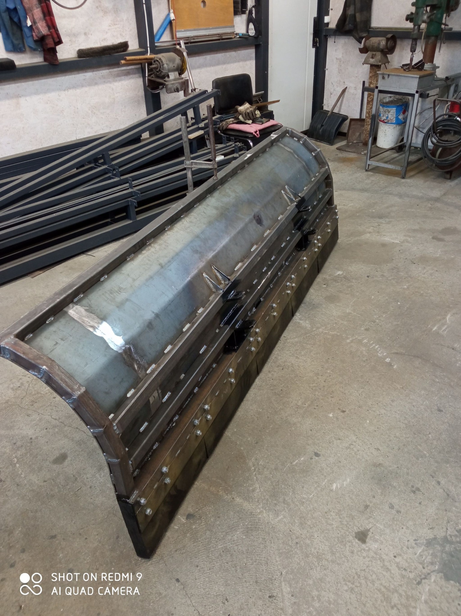 Metalowa konstrukcja z widocznymi spawami, prawdopodobnie element większej całości, leżąca na betonowej podłodze w warsztacie.