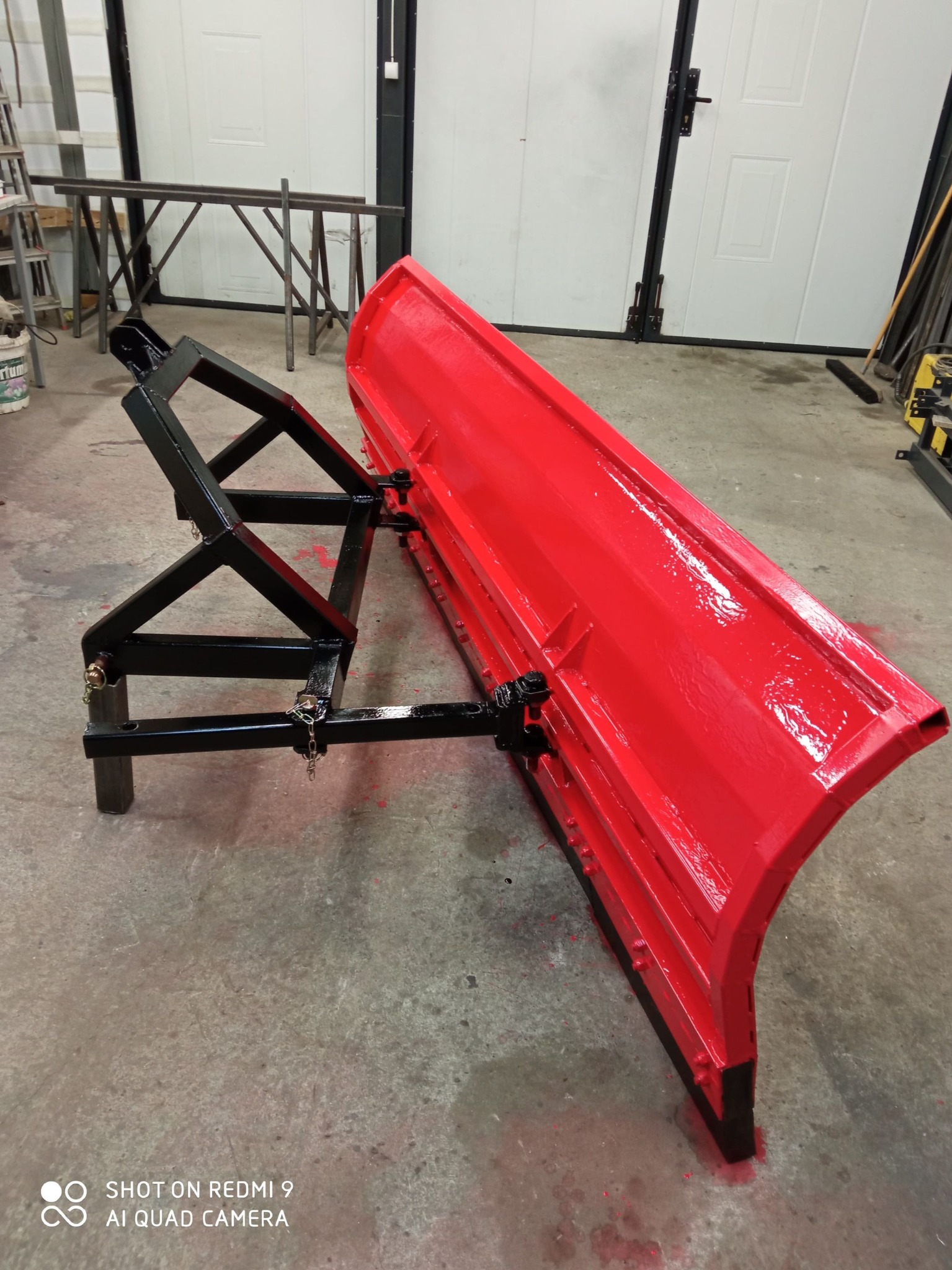 Czerwony pług śnieżny z czarną ramą, świeżo pomalowany, stoi na betonowej podłodze w warsztacie. Widoczne narzędzia i metalowe konstrukcje w tle.