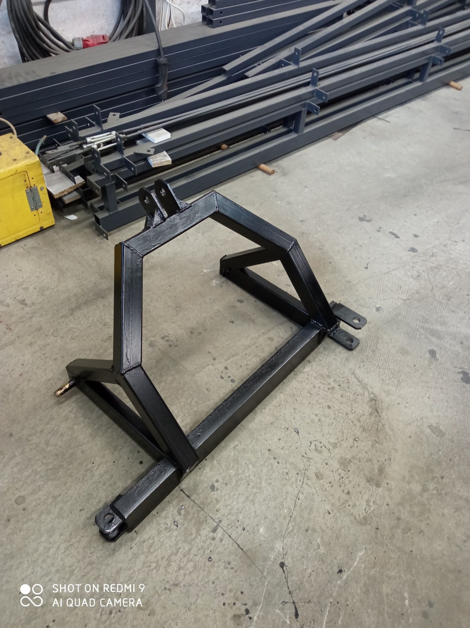 Czarna, spawana konstrukcja stalowa z profili zamkniętych, z widocznymi spawami, na betonowej posadzce warsztatu. W tle elementy konstrukcji stalowych i spawarka.