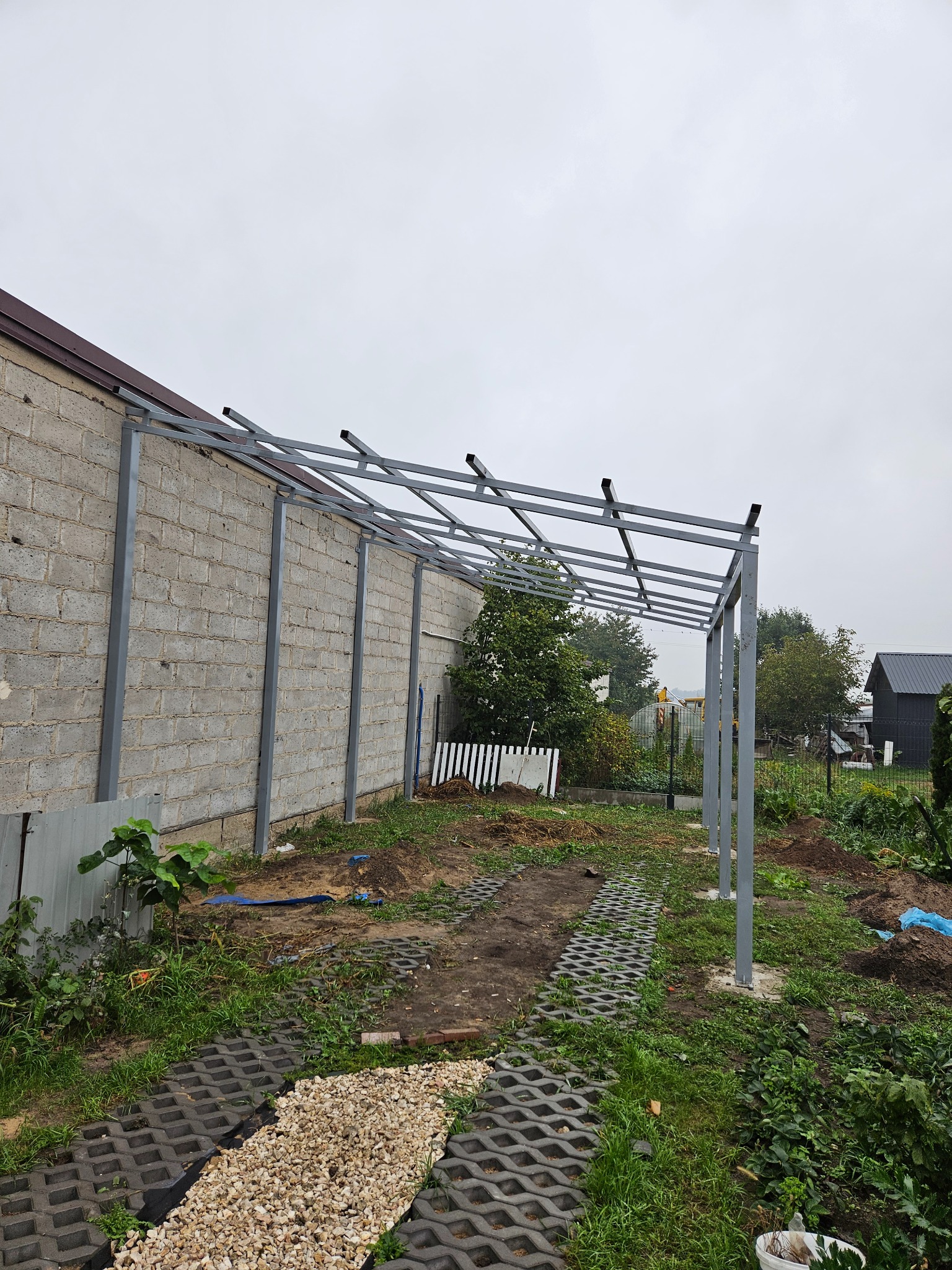 Stalowa konstrukcja z profili, prawdopodobnie zadaszenia, w trakcie montażu przy murowanym budynku. Widoczny fragment ścieżki z ażurowych płyt i kamieni.