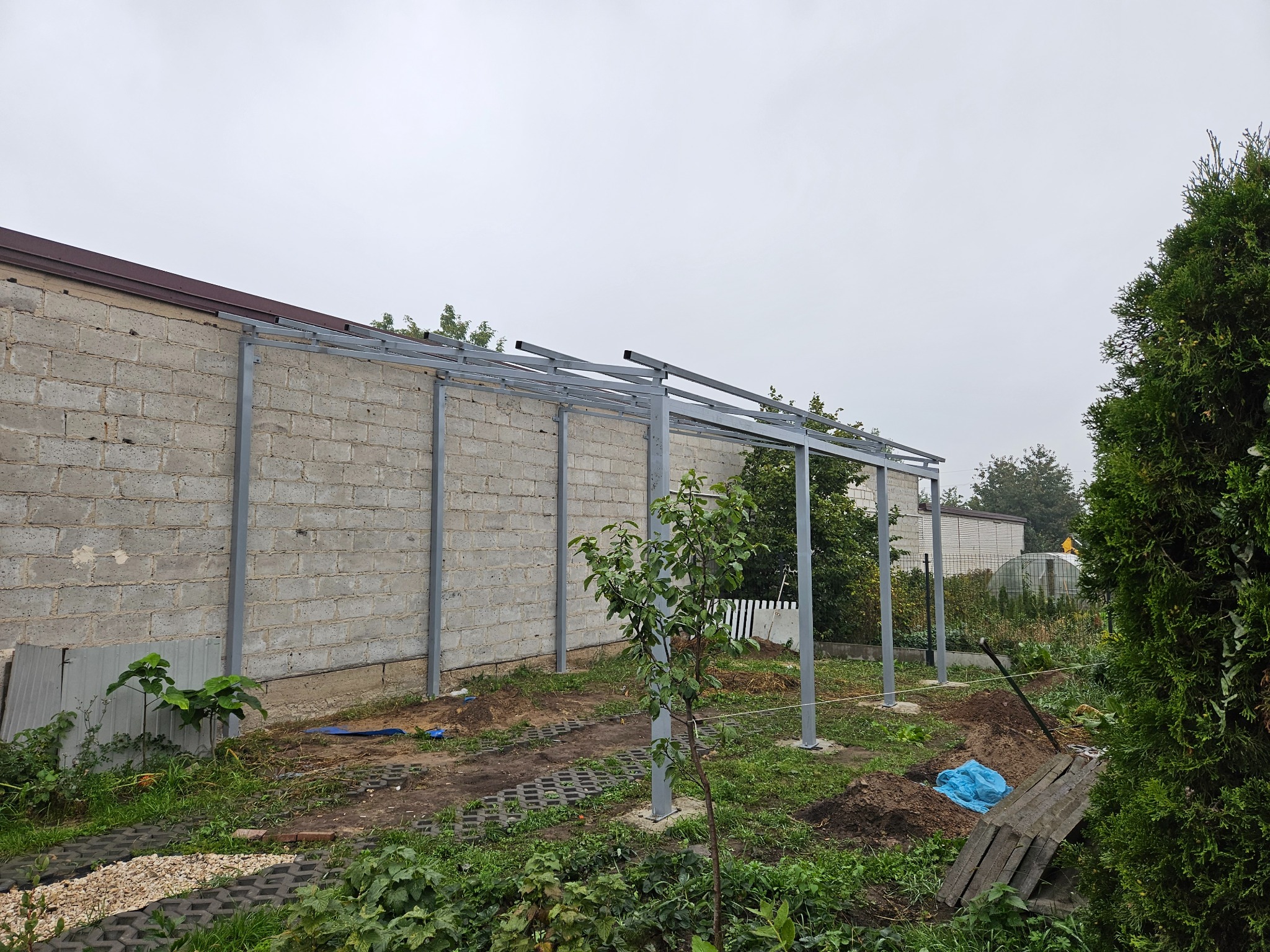 Stalowa konstrukcja z profili, prawdopodobnie zadaszenie tarasu lub werandy, w trakcie budowy przy murowanym budynku. Widoczne słupy i poziome elementy ramy.