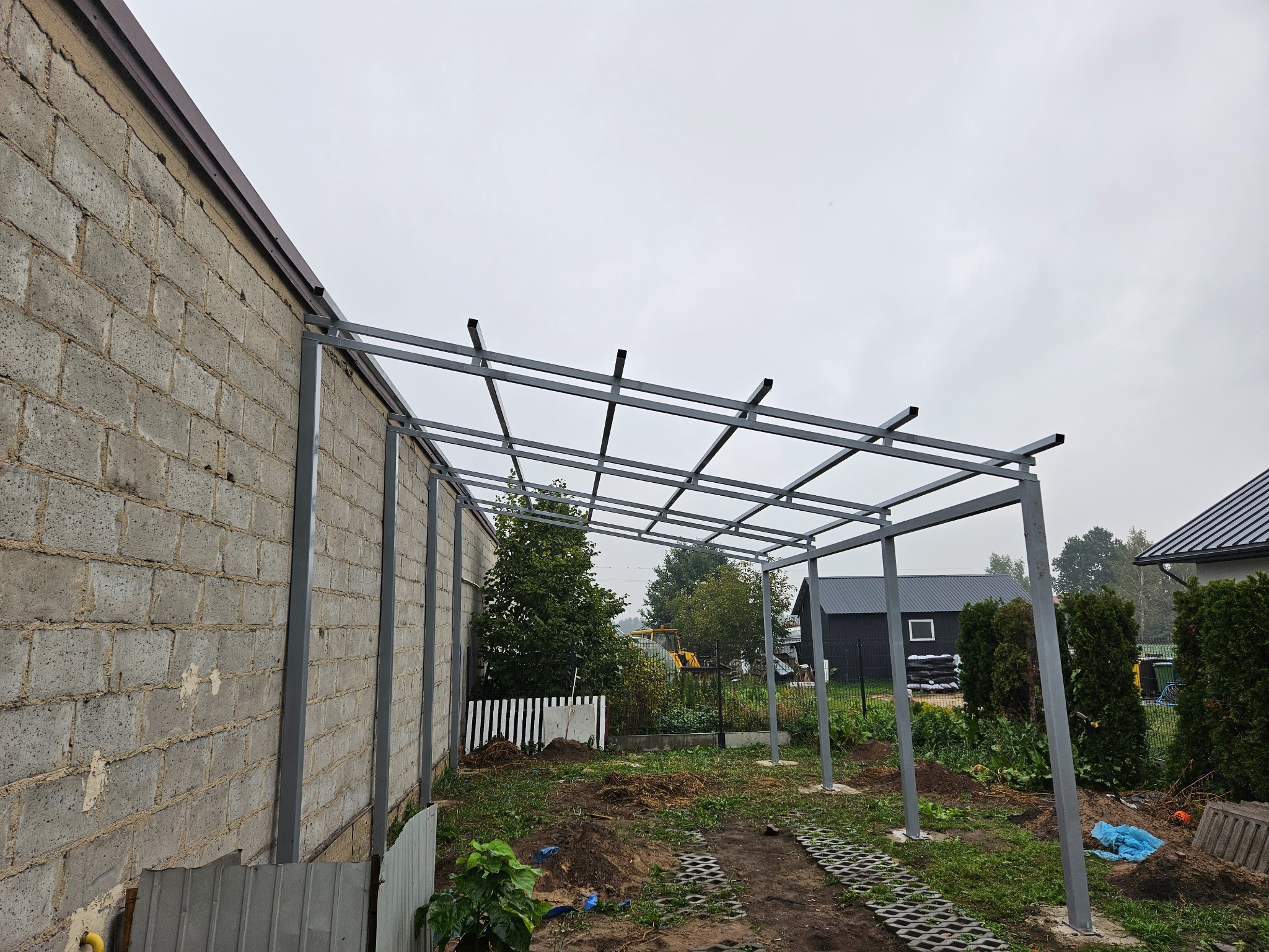 Metalowa konstrukcja z profili, prawdopodobnie zadaszenie tarasu, w trakcie budowy, widok z zewnątrz, na tle budynku z bloczków i zieleni.