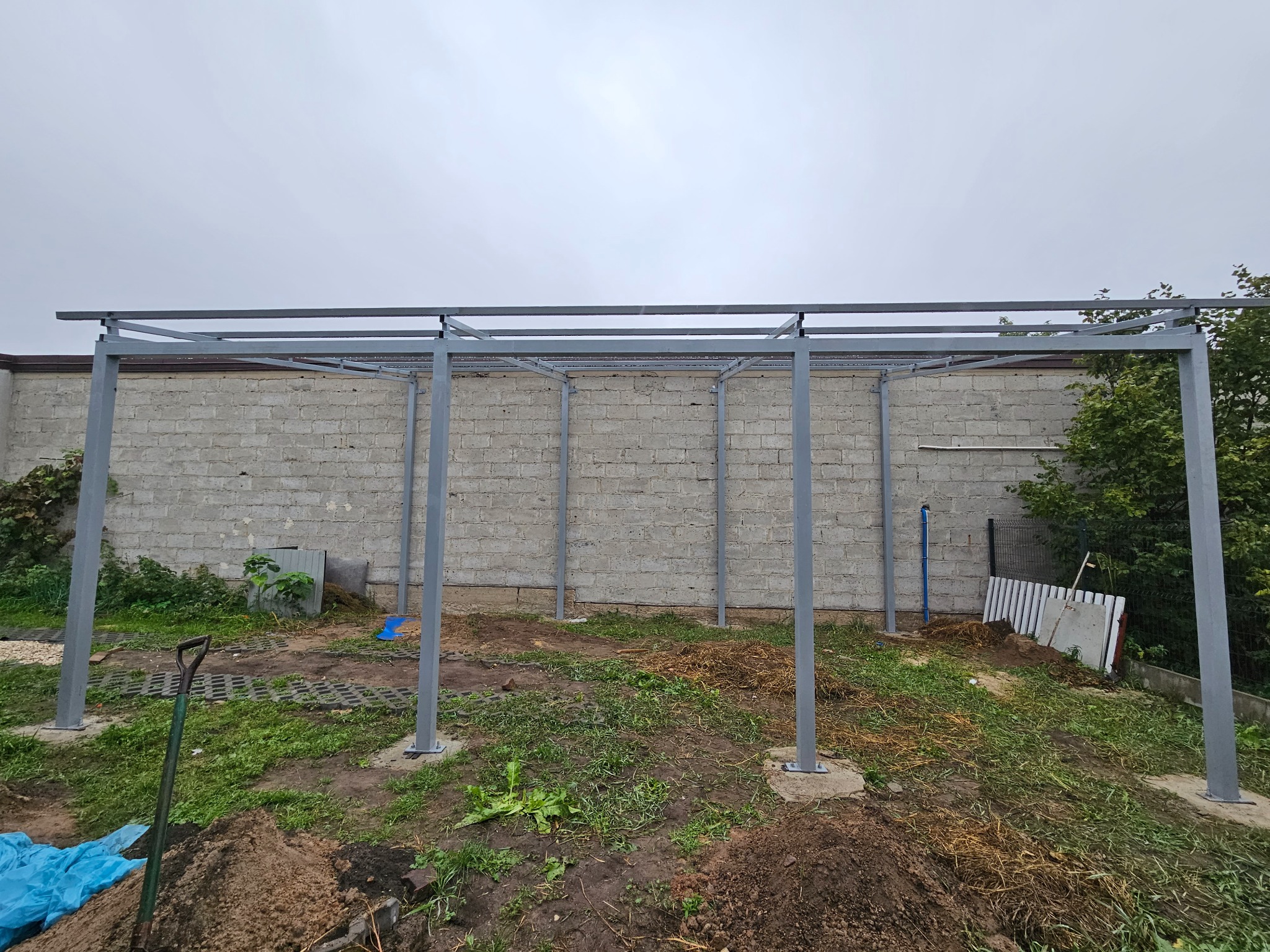 Szara, metalowa konstrukcja z profili, prawdopodobnie zadaszenia, w trakcie montażu na zewnątrz. Widoczne fundamenty, ziemia i ceglany mur w tle.
