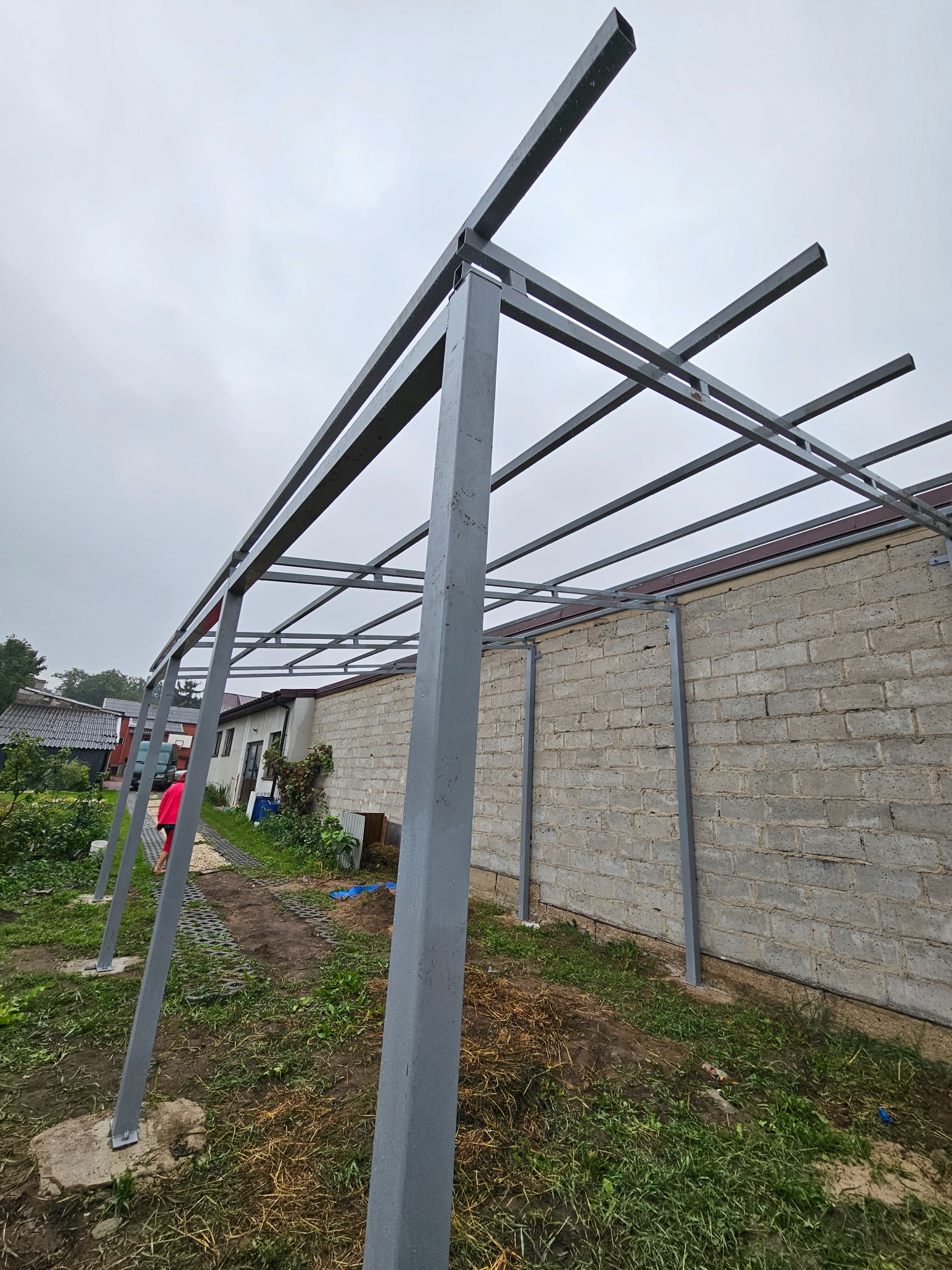 Szara, metalowa konstrukcja z profili kwadratowych, prawdopodobnie zadaszenie, w trakcie budowy. Widoczny fragment budynku z bloczków betonowych i niebo.
