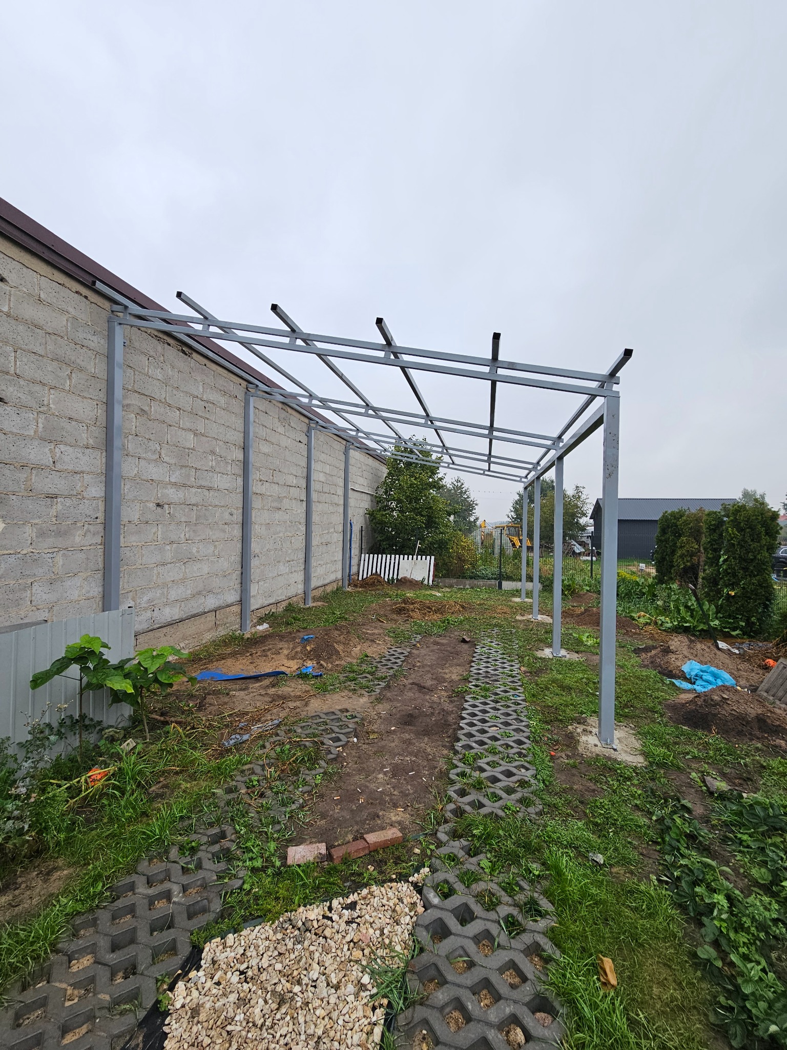 Szara, metalowa konstrukcja z profili, prawdopodobnie zadaszenia tarasu, w trakcie montażu przy budynku z szarej cegły. Widoczne elementy montażowe i otoczenie budowy.