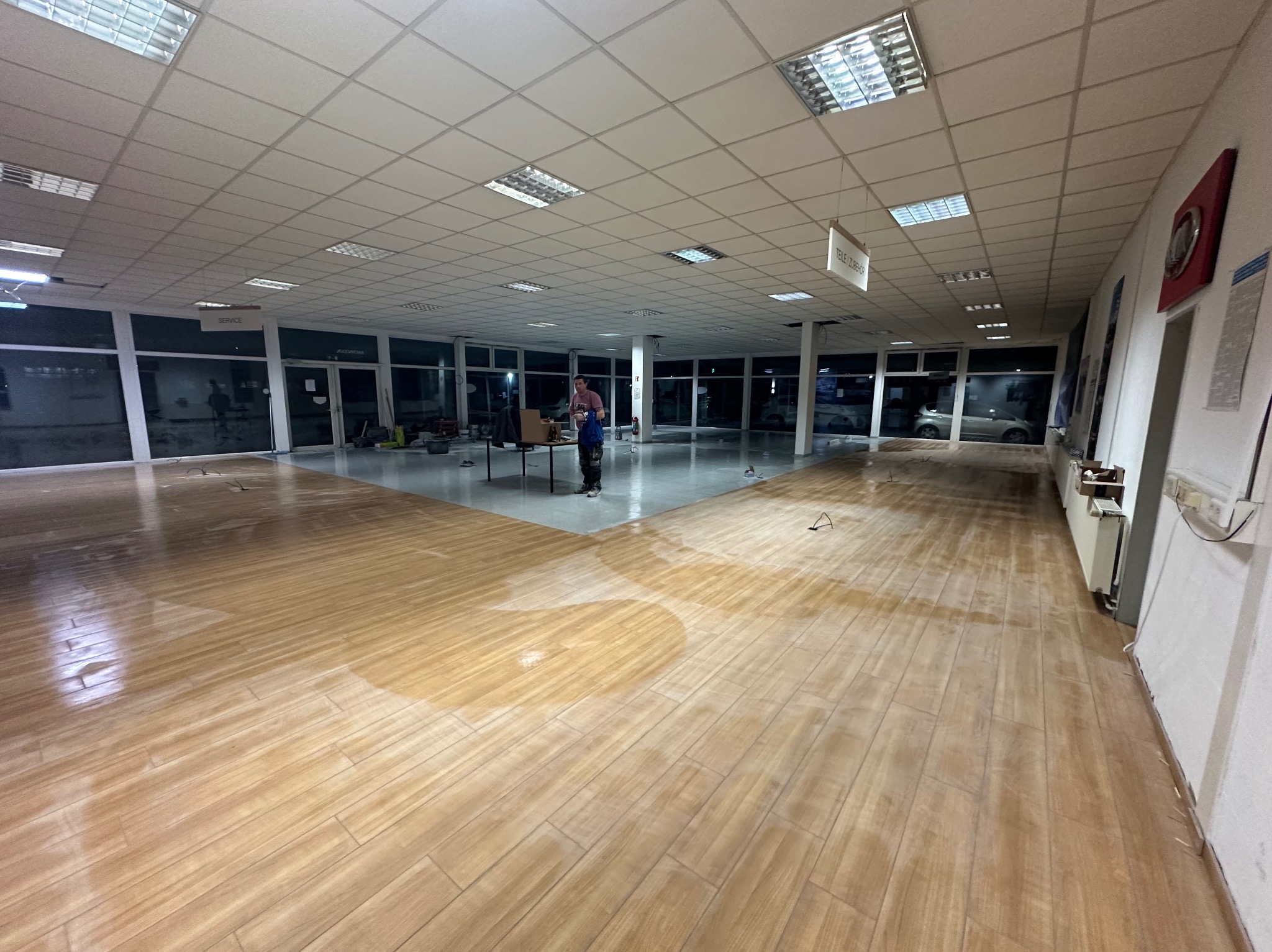 Układanie drewnopodobnych płytek podłogowych w przestronnym, jasnym salonie samochodowym. Widoczny pracownik i przygotowane stanowisko pracy.