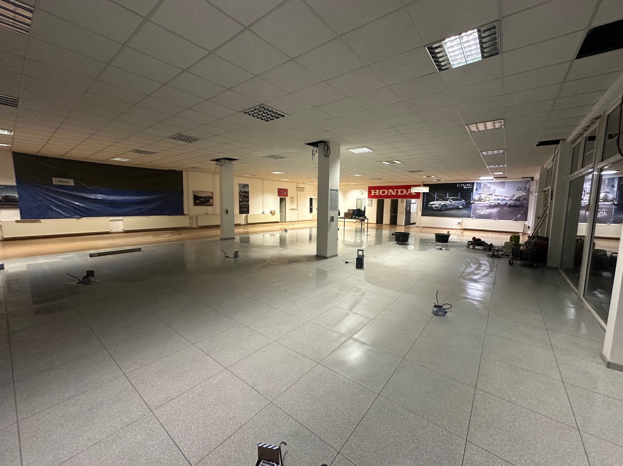 Jasne, przestronne wnętrze salonu samochodowego z nową, błyszczącą podłogą z płytek. Widoczne narzędzia i akcesoria do czyszczenia podłóg. Duże okna wpuszczają światło.