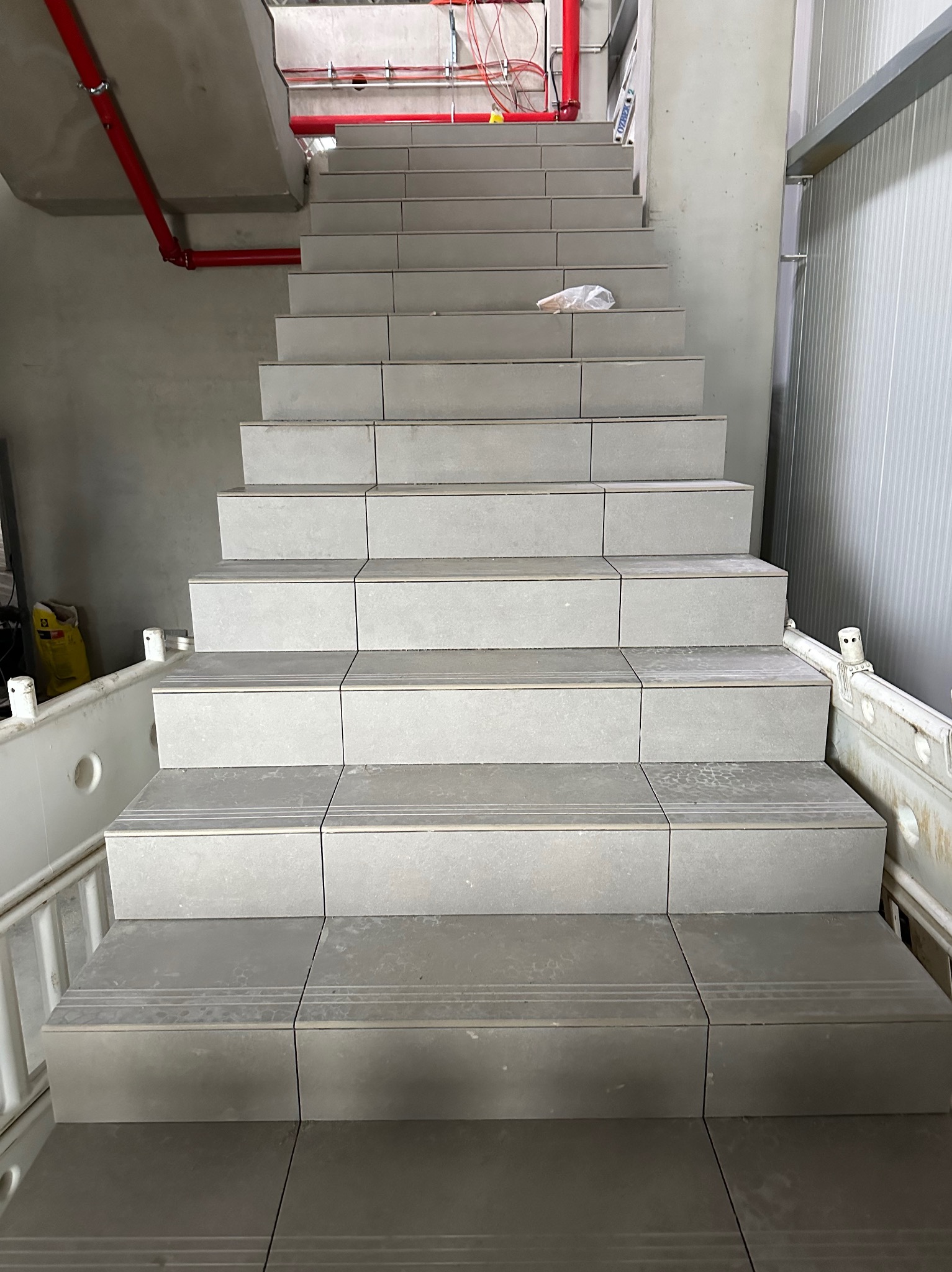 Wykończone schody z szarych płytek, widok z dołu. Na górze schodów leży worek foliowy. Czerwona rura na suficie. Widoczne ślady po pracy glazurniczej.