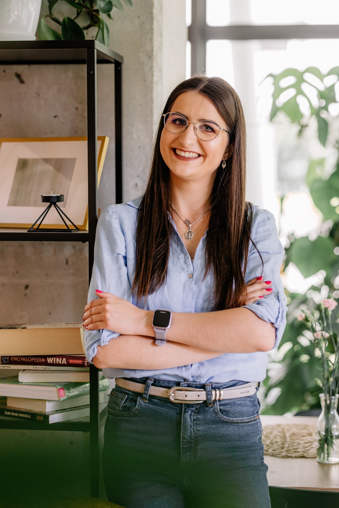 Uśmiechnięta brunetka w okularach, błękitnej koszuli i jeansach stoi na tle regału z książkami o winie i roślin doniczkowych, prezentując profesjonalny wizerunek.