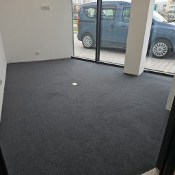 Perfect Floor Paweł Wojciechowicz - Wnętrze z nową, ciemnoszarą wykładziną dywanową. Widoczny fragment okna z zaparkowanym samochodem. Minimalistyczne wykończenie ścian i podłogi.