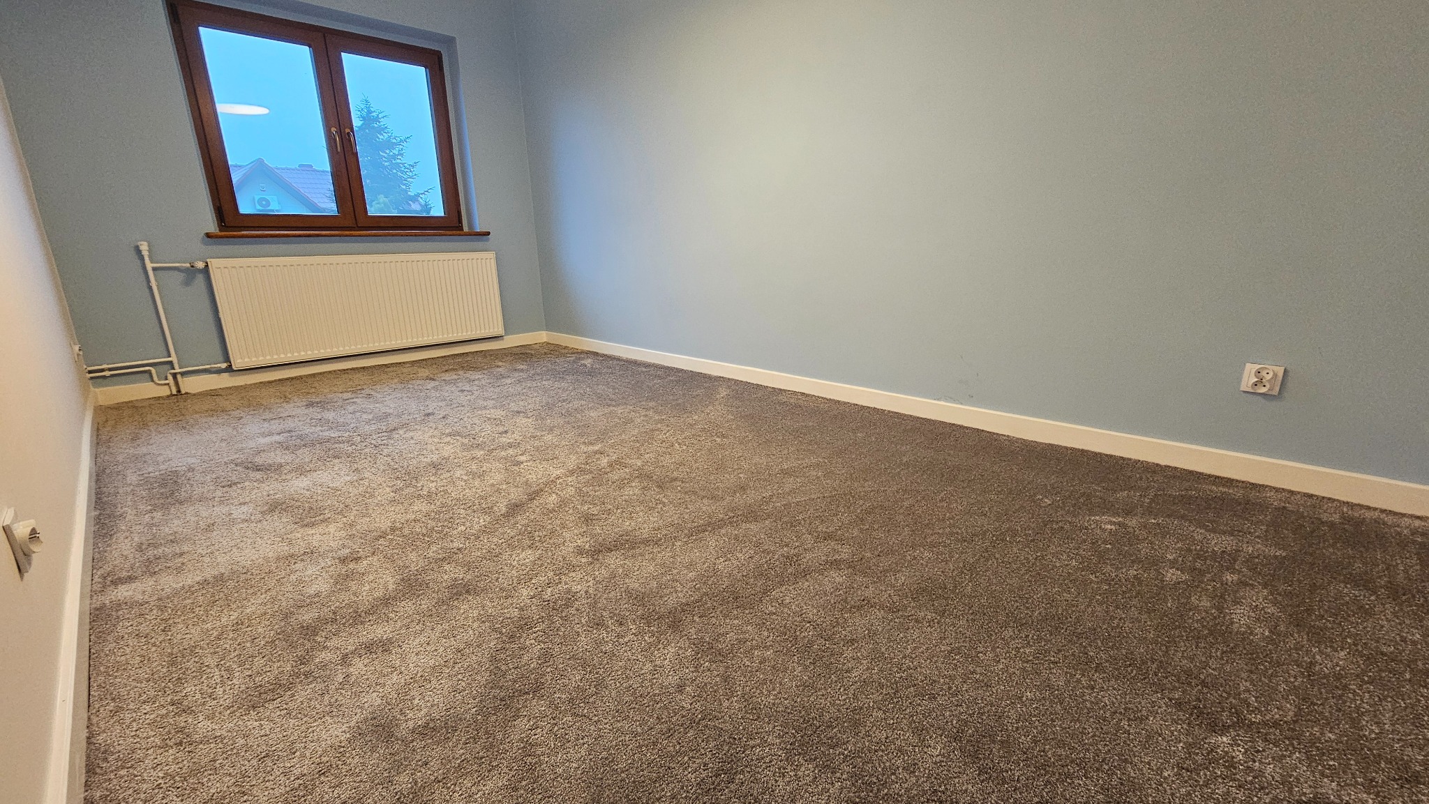 Pokój z nową, szarą wykładziną dywanową. Ściany pomalowane na jasny niebieski kolor. Widoczne okno i grzejnik. Listwy przypodłogowe w kolorze białym.