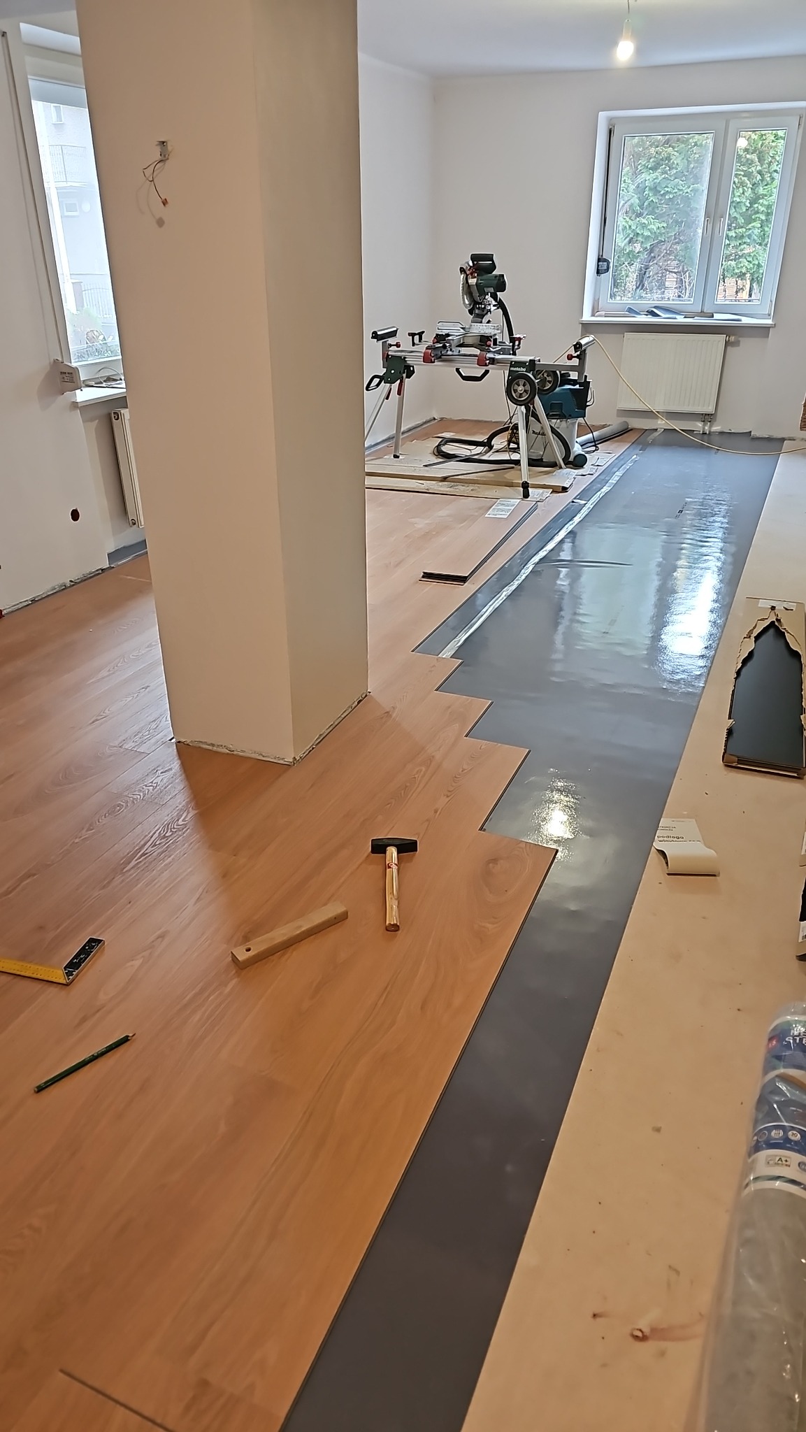 Układanie paneli podłogowych w jasnym wnętrzu. Widoczny fragment ułożonej podłogi, narzędzia pracy (młotek, miarka, ołówek) oraz maszyna do cięcia paneli.