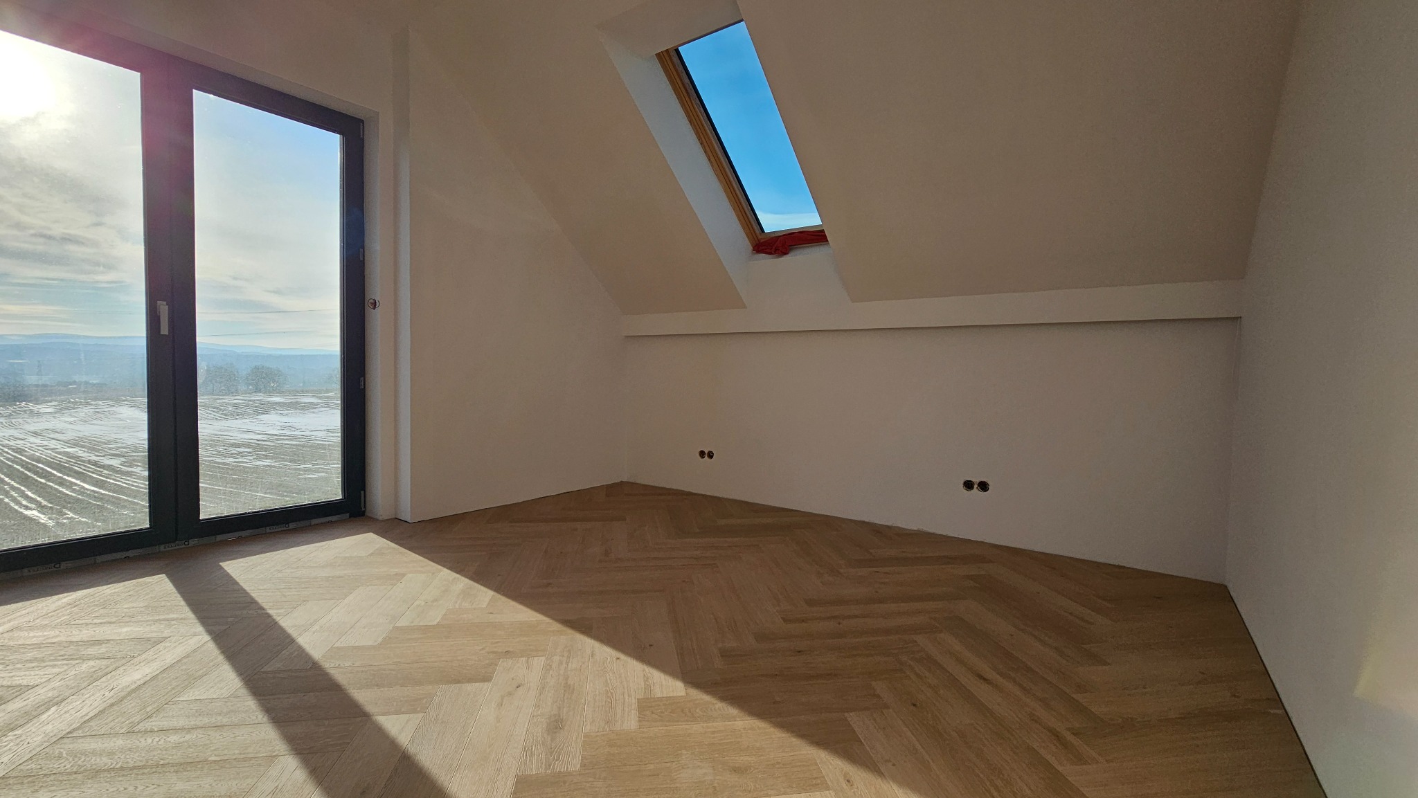 Jasne panele ułożone w jodełkę w pokoju na poddaszu z oknem dachowym i dużym oknem balkonowym z widokiem na zimowy krajobraz. Widoczne gniazdka elektryczne.