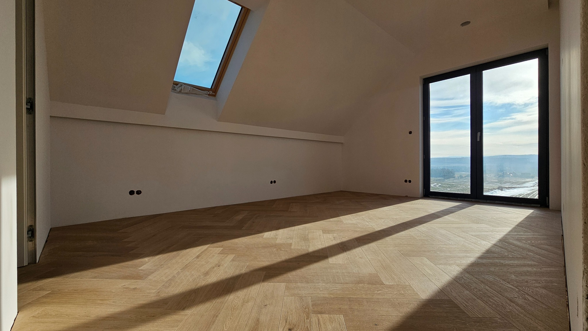 Jasne panele ułożone w jodełkę w pokoju na poddaszu z oknem dachowym i dużym oknem balkonowym z widokiem na krajobraz. Promienie słońca padają na podłogę.