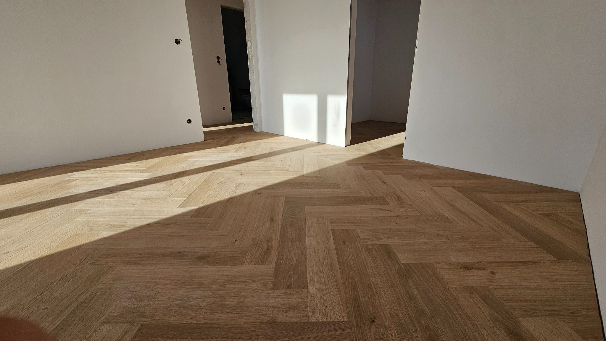 Ułożona parkietowa podłoga w jodełkę, oświetlona promieniami słońca wpadającymi przez otwarte drzwi w jasnym wnętrzu. Widoczne gniazdka elektryczne na ścianie.