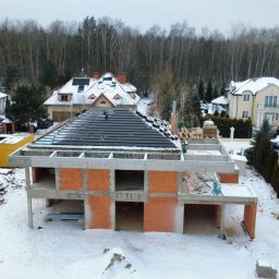 Budowa domu z czerwonej cegły w zimowej scenerii. Częściowo pokryty dach z czarnej dachówki. W tle inne domy i las. Widok z góry.