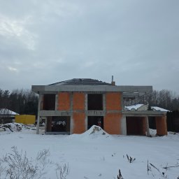 Surowy stan domu z czerwonej cegły w zimowej scenerii. Widoczne elementy konstrukcyjne i fundamenty. Na pierwszym planie śnieg i roślinność.