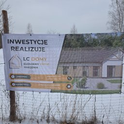 Baner reklamowy firmy budowlanej LC Domy z wizualizacją domu murowanego, umieszczony na ogrodzeniu z siatki w zimowym krajobrazie. Widoczne dane kontaktowe i hasło: Budujemy Twoje Marzenia.