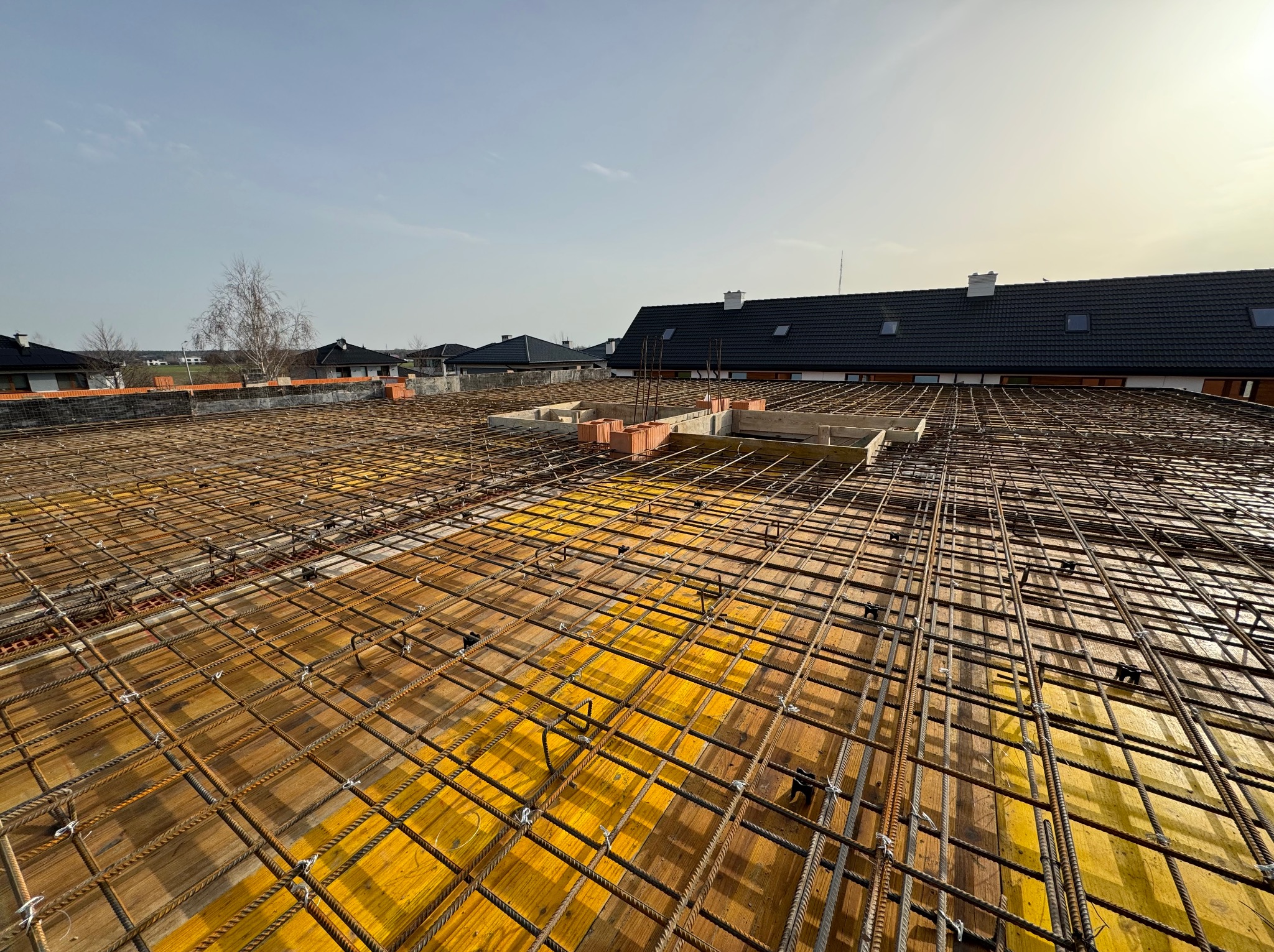 Zbrojenie stropu na deskowaniu z żółtej sklejki, widok z góry na budowę domu jednorodzinnego, w tle zabudowa mieszkalna i błękitne niebo.