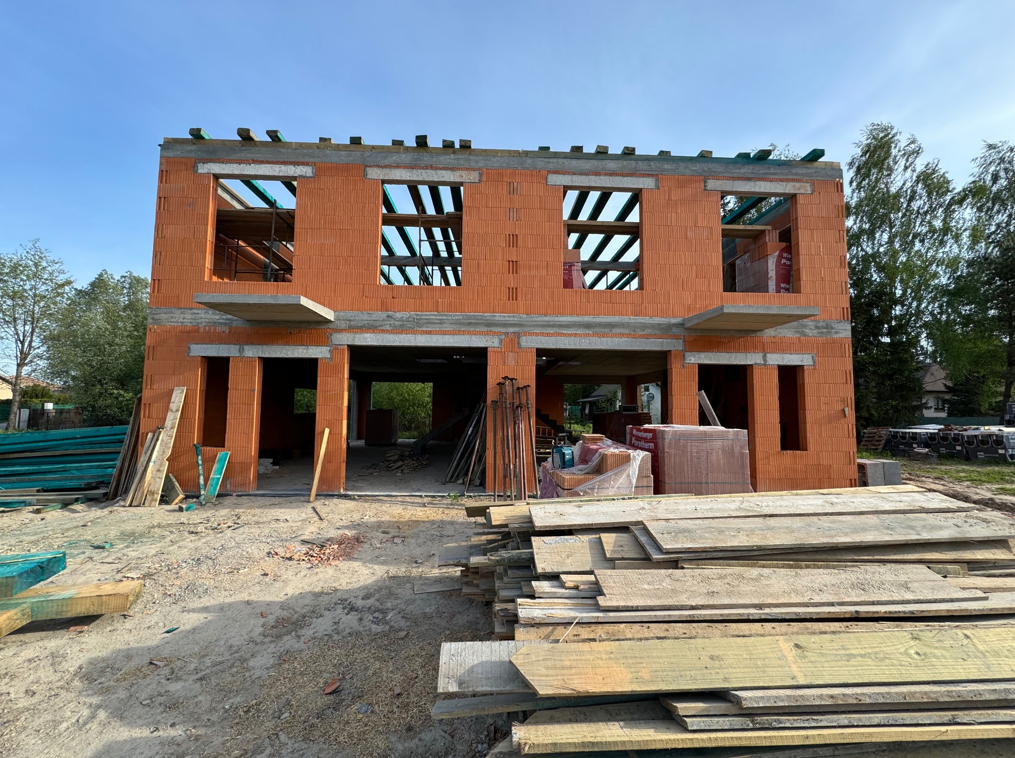 Dwukondygnacyjny dom murowany w trakcie budowy, widoczne pustaki ceramiczne Porotherm, otwory okienne i drzwiowe, betonowe nadproża, drewniane stemple i deski szalunkowe na placu budowy.