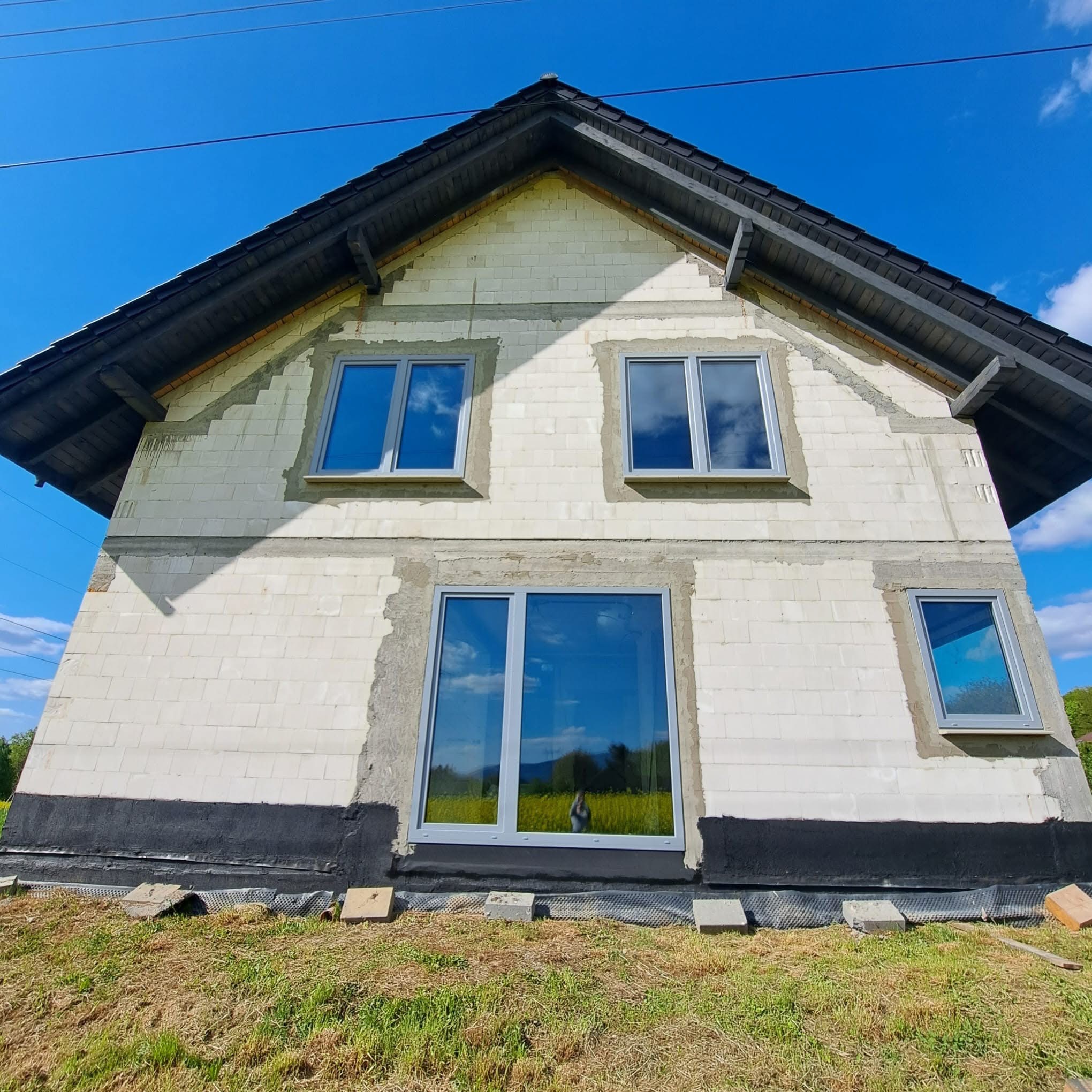 Surowy stan domu murowanego z widocznymi oknami i izolacją fundamentów na tle błękitnego nieba. Widok z dołu, podkreślający etap budowy.