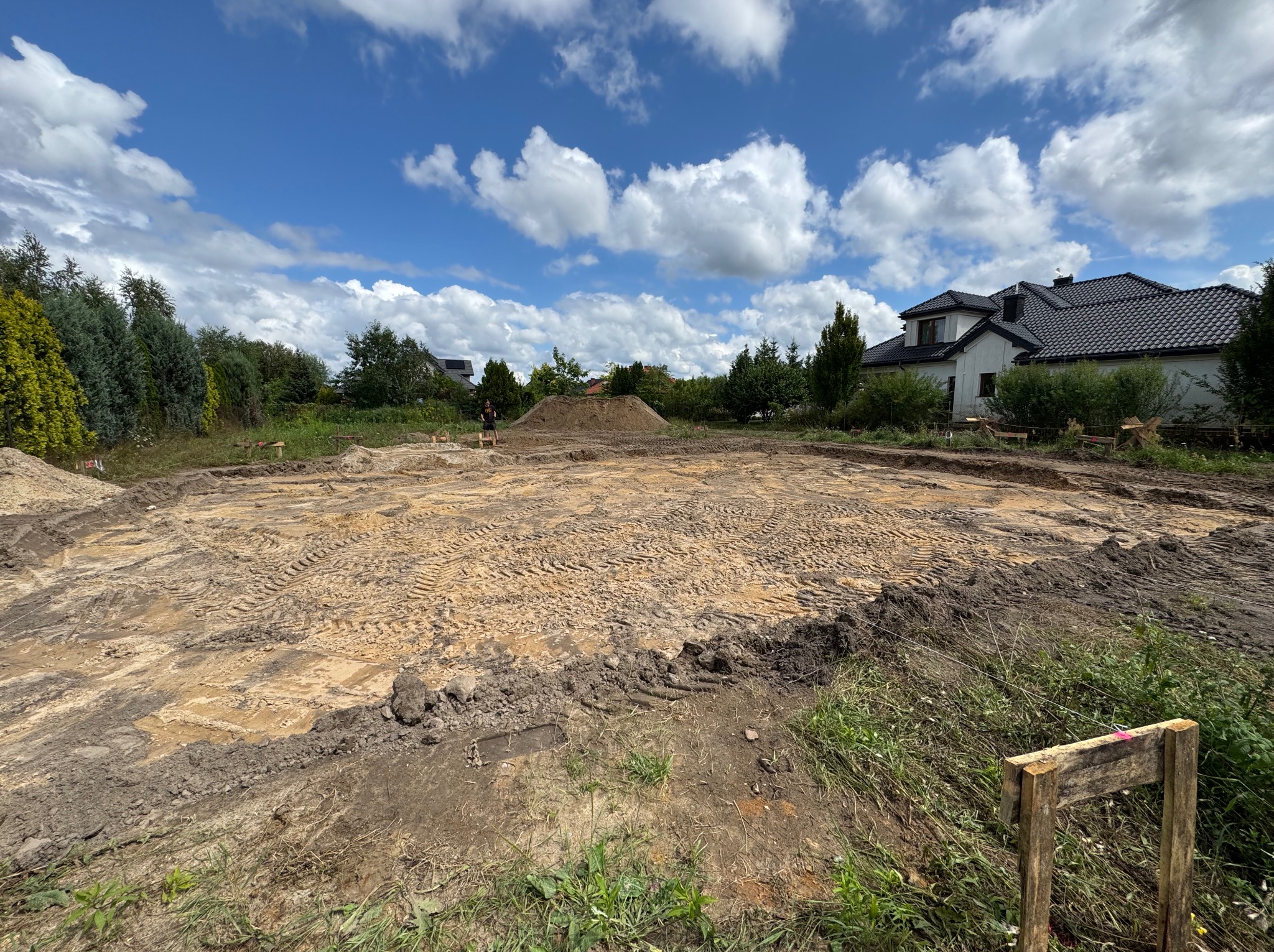 Przygotowany teren pod budowę domu murowanego. Widoczny wyrównany plac z piaskiem, ślady opon, w tle częściowo widoczny dom i błękitne niebo z chmurami.