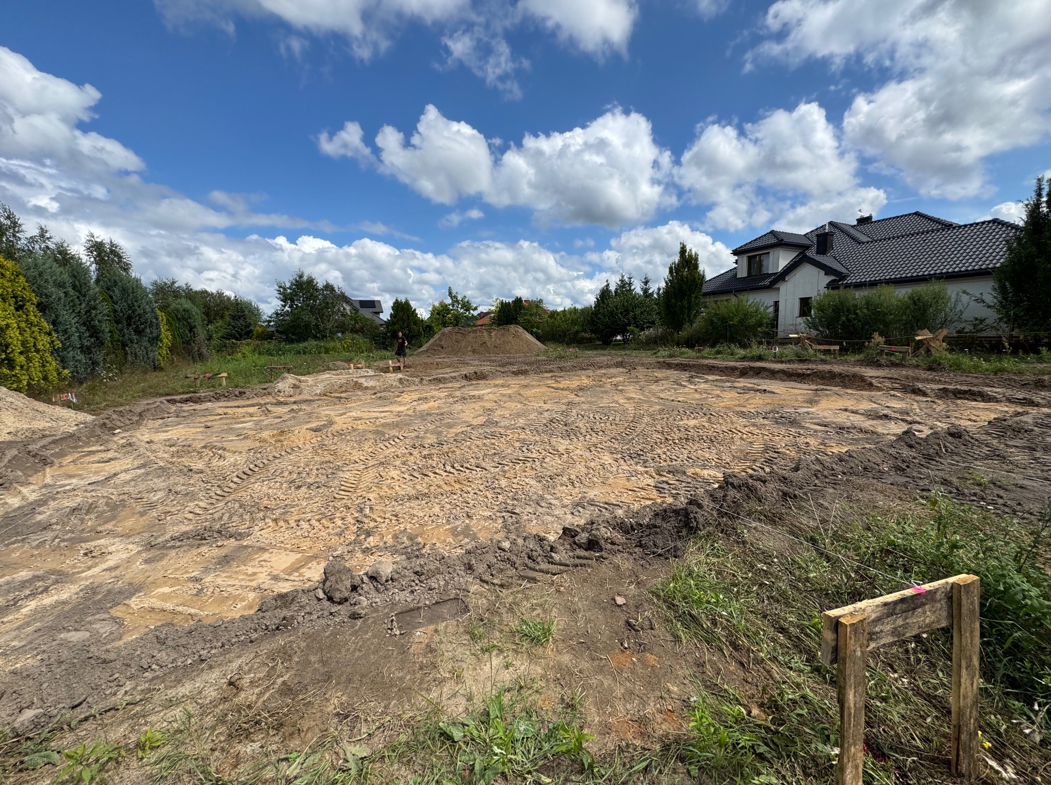 Przygotowany teren pod budowę domu. Widoczne wytyczone fundamenty, wyrównany grunt i hałda ziemi w tle. W oddali dom z ciemnym dachem i błękitne niebo z chmurami.