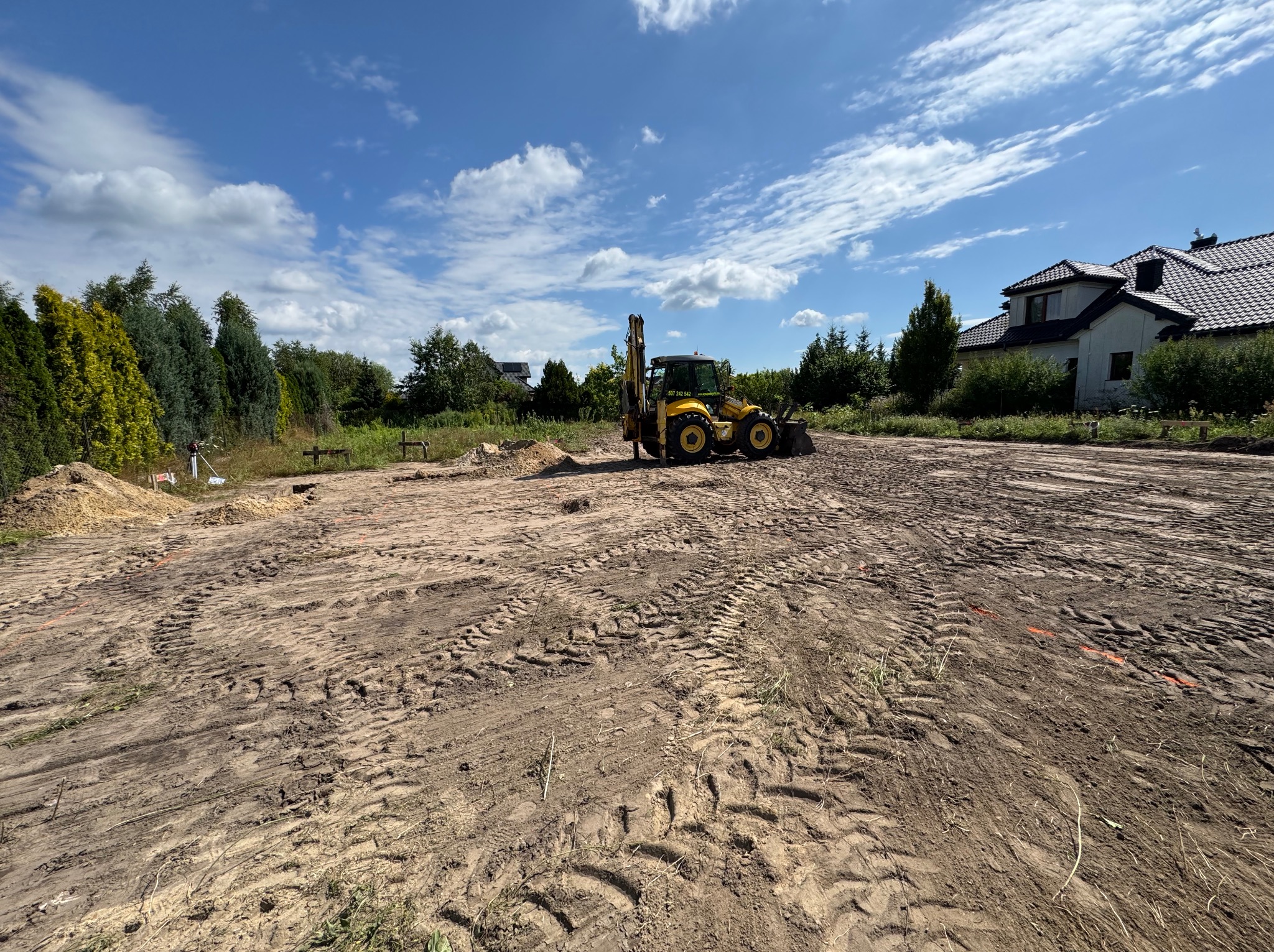 Żółta koparko-ładowarka na przygotowanym terenie budowy pod dom murowany, z widocznymi śladami opon i składowiskami ziemi. W tle dom i drzewa.