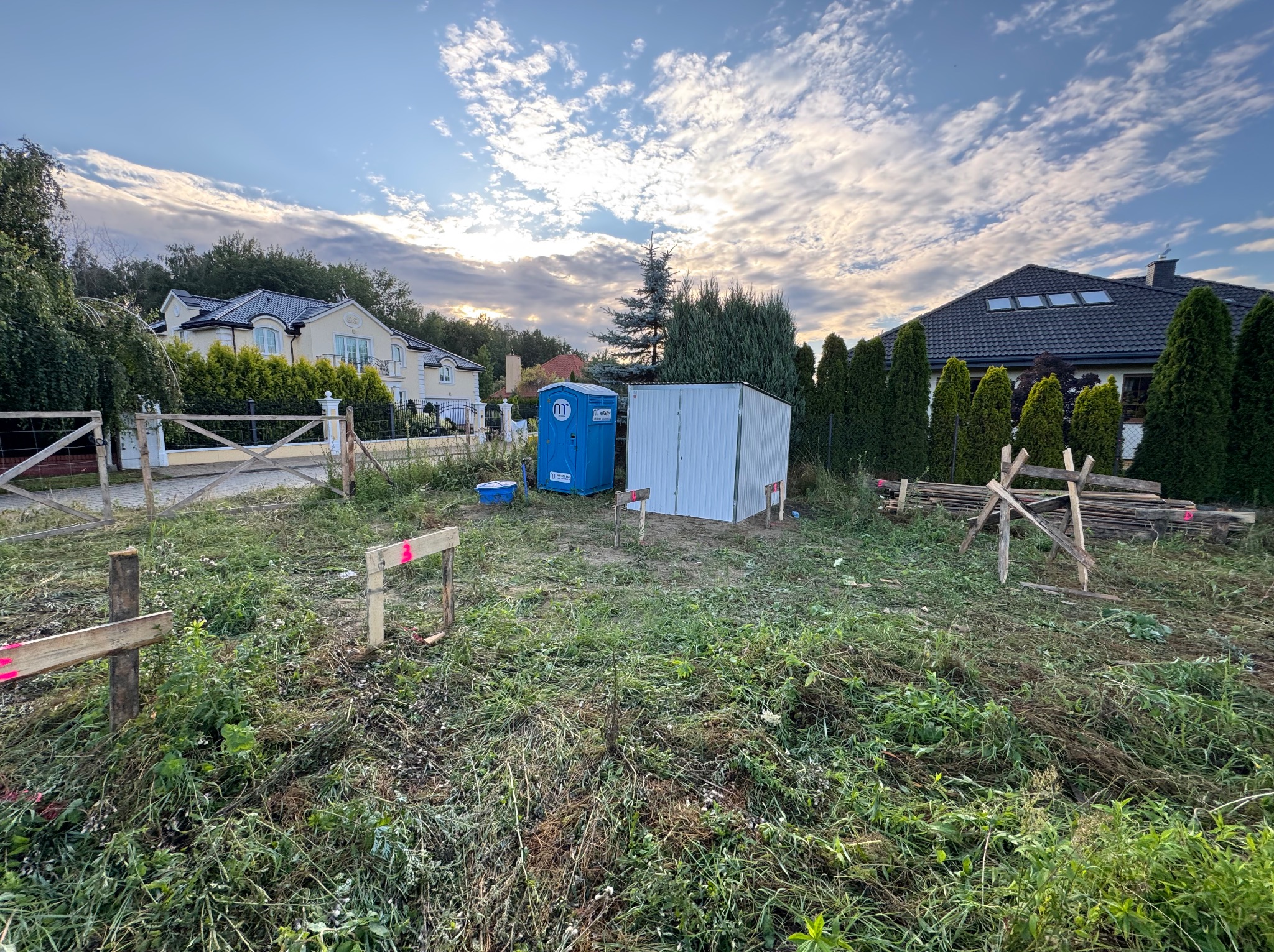 Działka budowlana z wyznaczonym obrysem przyszłego domu. Widoczne drewniane słupki i deski, przenośna toaleta i składzik na narzędzia. W tle domy jednorodzinne.