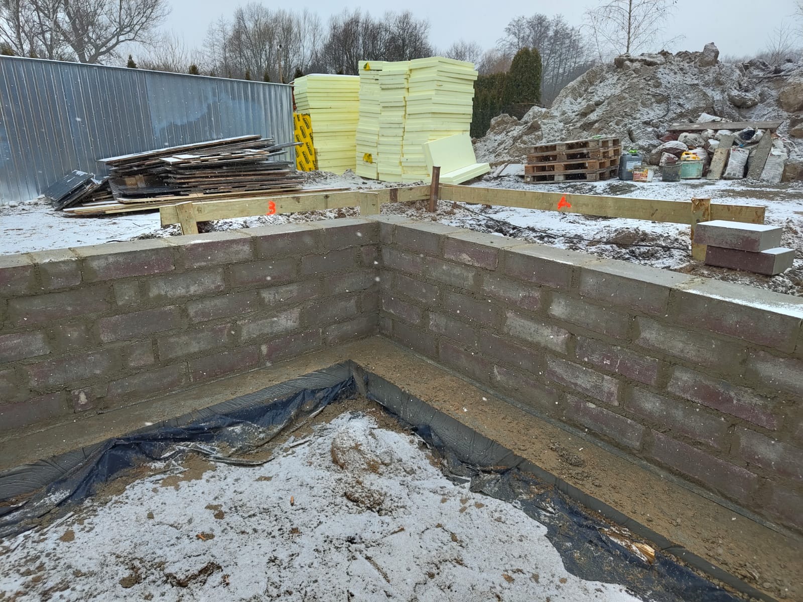 Wykop fundamentów pod dom z częściowo wymurowanymi ścianami z cegły. W tle materiały budowlane i ogrodzenie. Zimowa sceneria z padającym śniegiem.