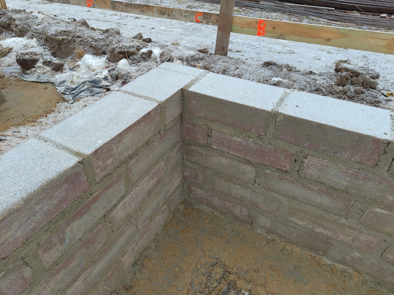 Wznoszenie muru z czerwonej cegły i szarego bloczka betonowego na fundamencie. Widoczne drewniane stemple i zbrojenie. Prace murarskie w toku, surowy stan.