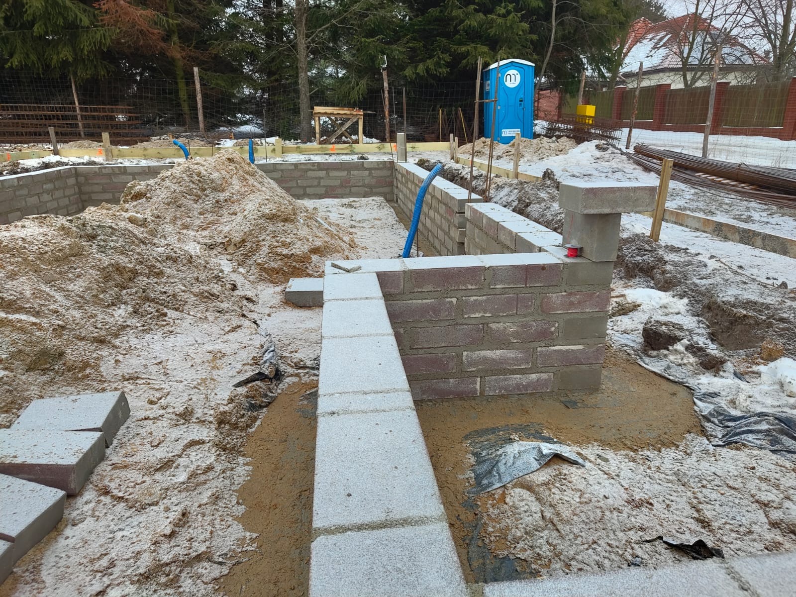 Fundamenty domu z cegły, częściowo pokryte śniegiem, z widoczną niebieską rurą i przenośną toaletą w tle. Etap budowy w zimowej scenerii.